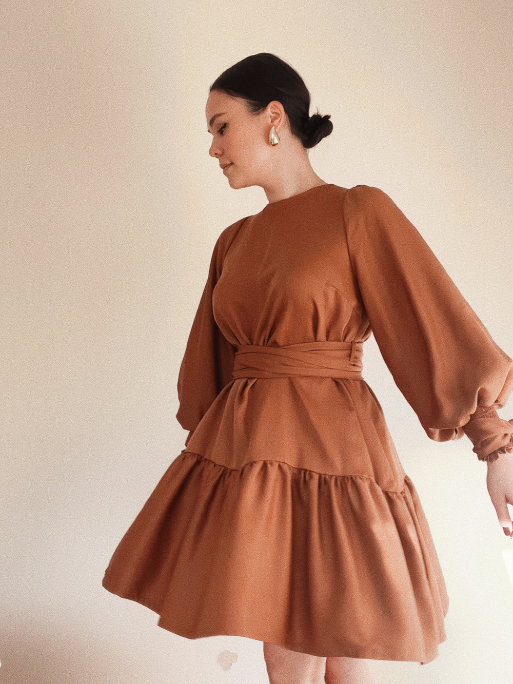 Woman wearing the Audrey Dress sewing pattern from Veronica Tucker on The Fold Line. A dress pattern made in linen or compact cotton fabric, featuring a voluminous sleeve with a shirred cuff and ties, a round neck, double wrap waist tie, gathered hem panel, and above-knee length.