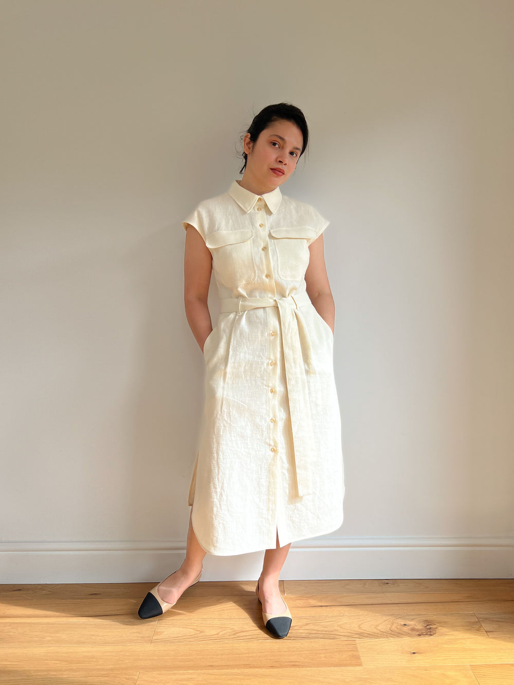 Woman wearing the Aurea Dress sewing pattern from Bella Loves Patterns on The Fold Line. A sleeveless shirt dress pattern made in linen or cotton fabric, featuring dropped shoulders, a back yoke, pointed shirt collar, collar stand, centre front button placket, front patch pockets with flaps, tie belt, in-seam side pockets, side slits, a curved hem, and midi length.