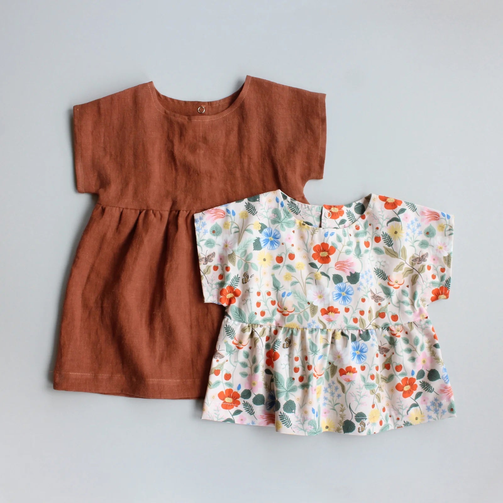 The Baby/Child Nora Dress and Top sewing pattern from Oh Me Oh My Sewing on The Fold Line. A dress or top pattern made in quilting cotton, washed linen, double gauze, cotton lawn, voile, or rayon challis fabric, featuring an oversized fit, round neckline, grown-on short sleeves, snaps on the back bodice, and a gathered skirt or peplum.