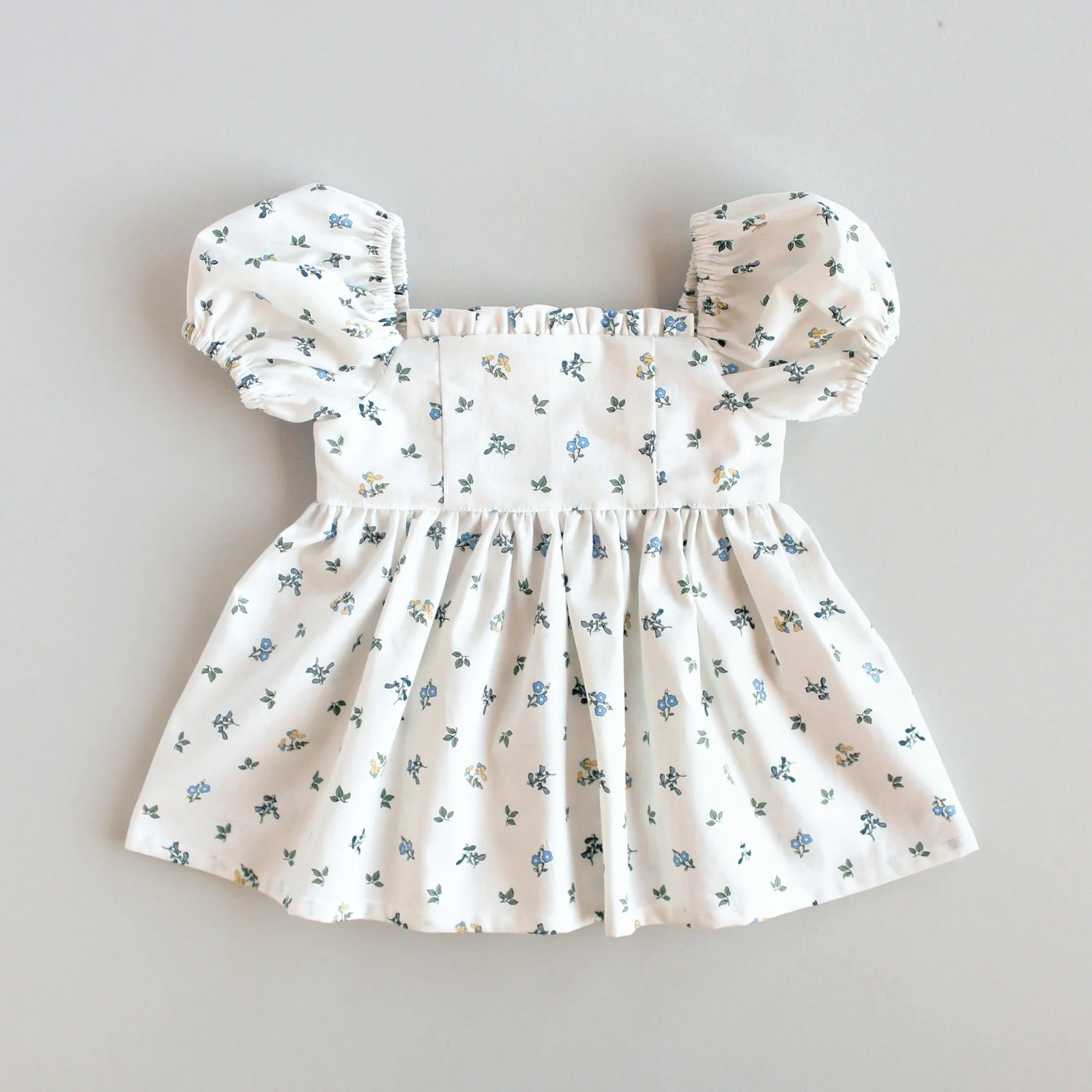 The Baby/Child Sophie Dress sewing pattern from Oh Me Oh My Sewing on The Fold Line. A dress pattern made in quilting cotton, washed linen, cotton lawn, voile, or rayon challis fabric, featuring a square neckline with a ruffle, empire waist, short puff sleeves, gathered skirt, and button closure at the back.