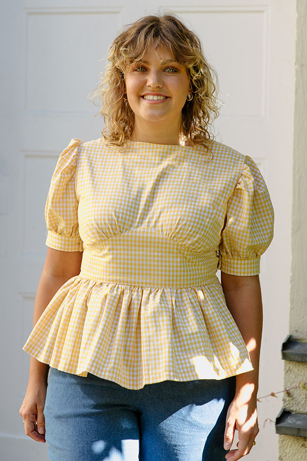 Woman wearing the Bella Peplum Blouse sewing pattern from JULIANA MARTEJEVS on The Fold Line. A top pattern made in cotton fabric, featuring short puff sleeves, a fitted waist, gathered peplum, and bow at the back.