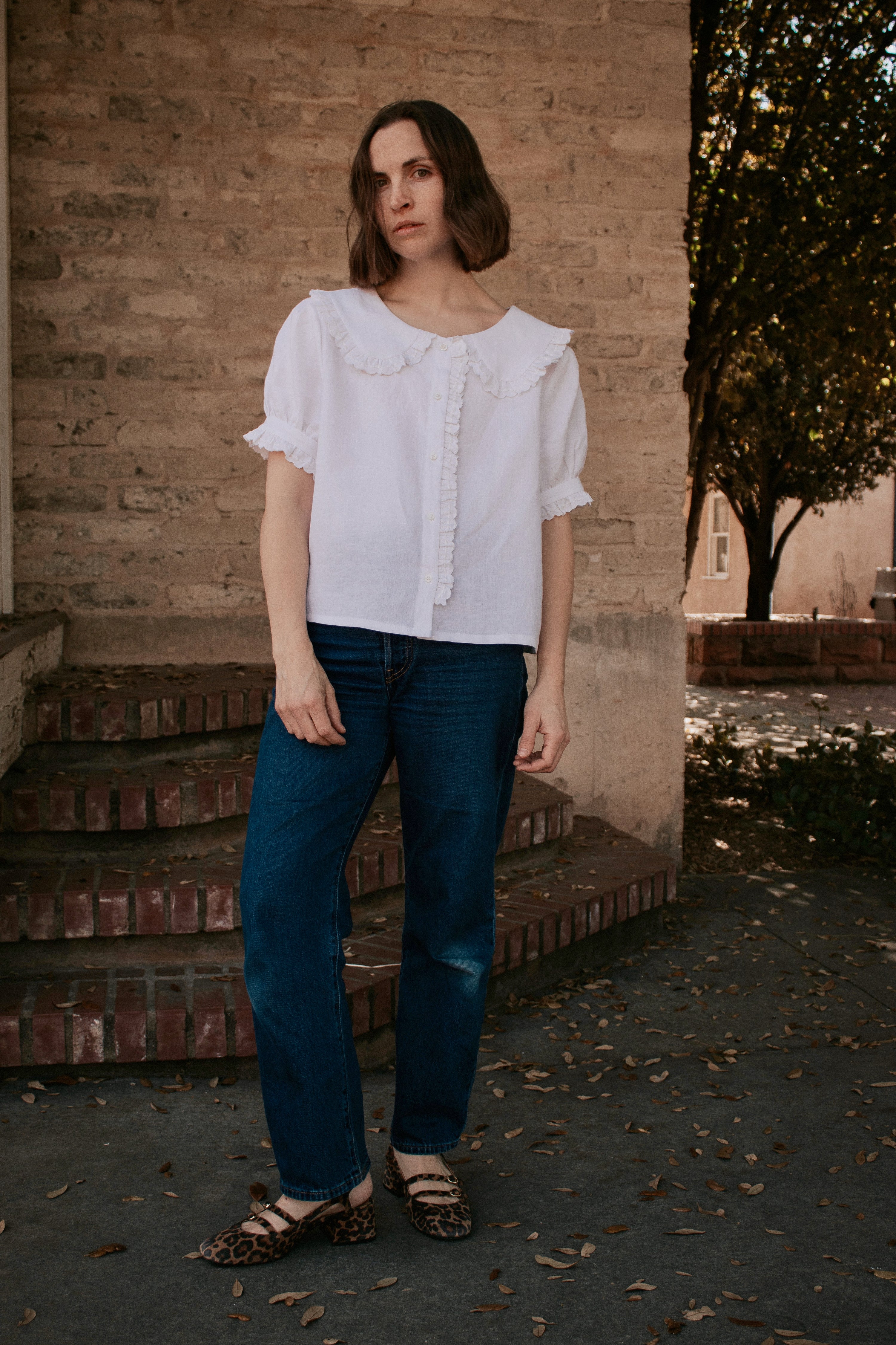 Woman wearing the Beth Blouse sewing pattern from Madswick on The Fold Line. A top pattern made in linen, viscose, tencel, chambray, cotton lawn, cotton poplin, silk, or cotton twill fabric, featuring a Peter Pan collar, front button placket, and short puff sleeves, all with ruffles.