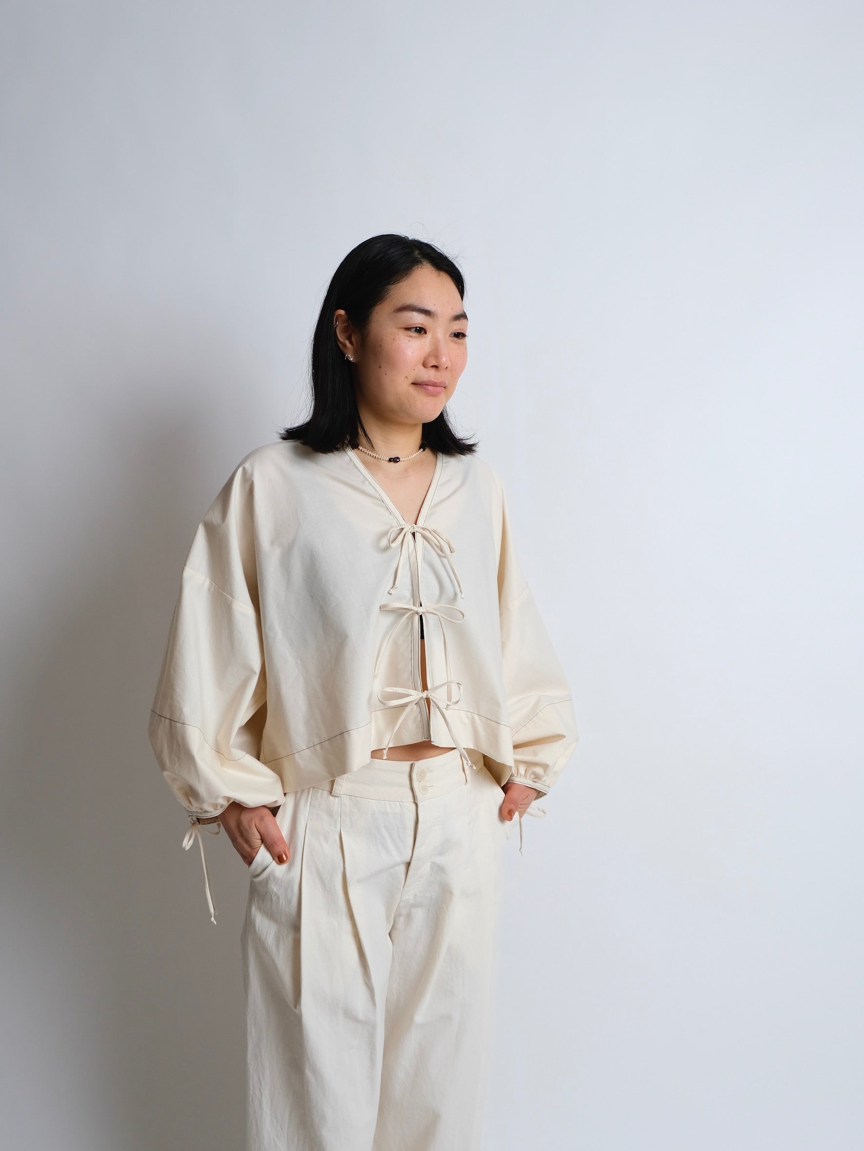 Woman wearing the Bow Top Hack sewing pattern from Birgitta Helmersson on The Fold Line. An oversized top pattern made in cotton or linen fabric, featuring a V-neckline, tie front, and voluminous long sleeves with ties.