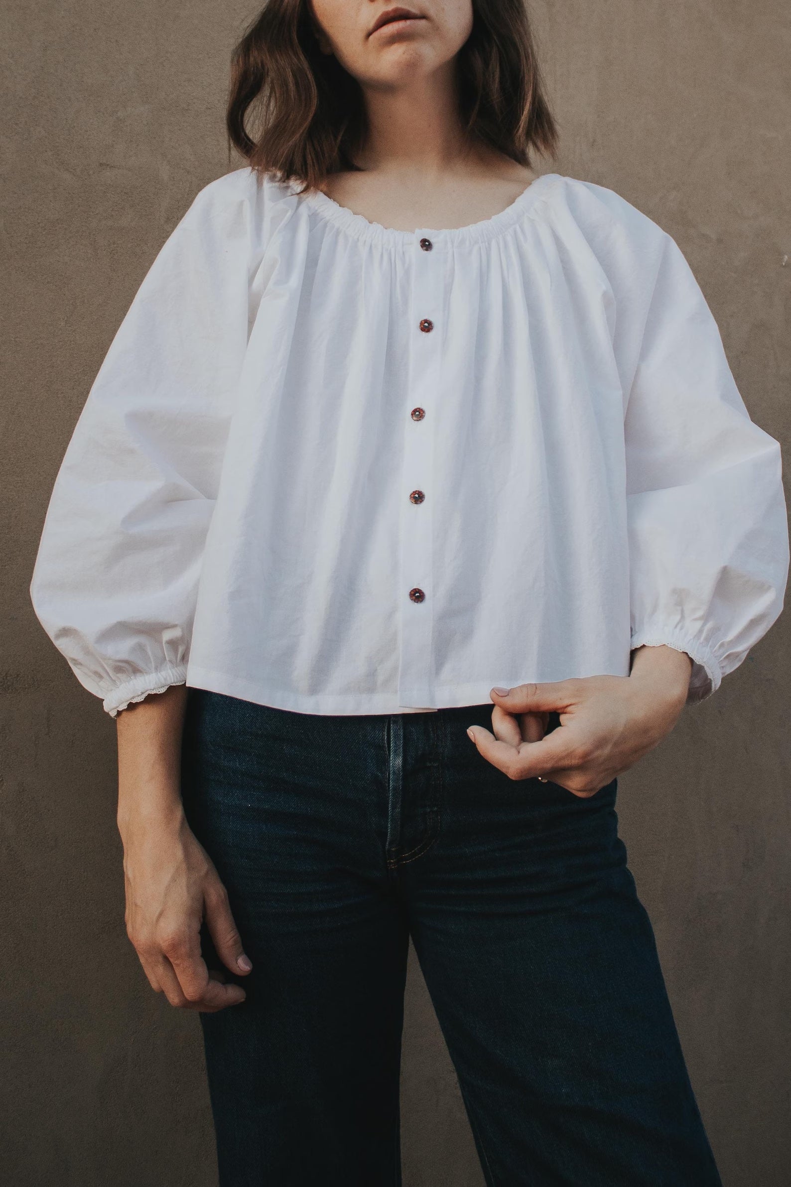 Woman wearing the Carol Blouse sewing pattern from Madswick on The Fold Line. A top pattern made in rayon, tencel, cotton voile, cotton lawn, poplin, or linen fabric, featuring a relaxed fit, gathered scoop neckline, button front, and 3/4 length raglan sleeves.