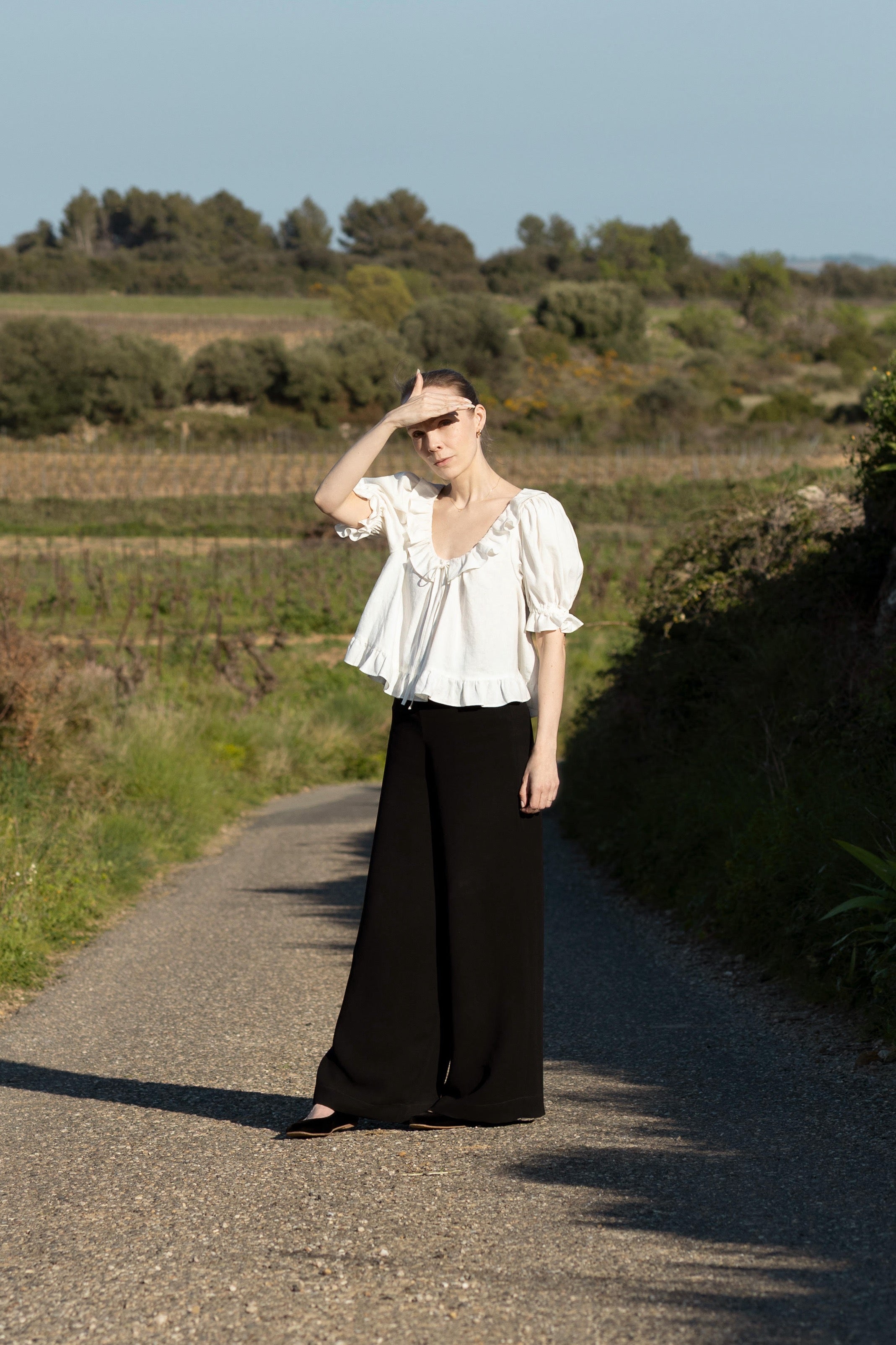 Woman wearing the Celine Blouse sewing pattern from Silversaga Patterns on The Fold Line. A top pattern made in cotton lawn, voile, lightweight linen, or soft poplin fabric, featuring a relaxed fit, deep round neckline with a ruffle and tie, gathers at the front and back, short puffed sleeves, and a ruffle at the hem.