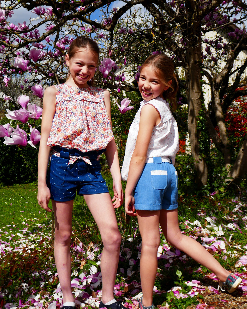 Girls wearing the Child/Teen Xylo Shorts sewing pattern from Petits D'om on The Fold Line. A shorts pattern made in denim, cotton, light gabardine, twill, double gauze, or chambray fabric, featuring an elasticated waist, mock fly, optional waistband frill, wide belt loops with a tie belt, and pockets.