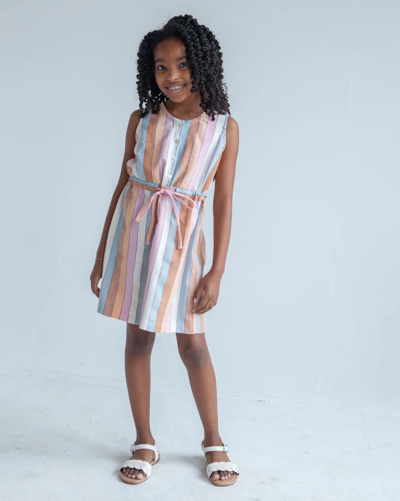 Girl wearing the Children's Mini Southport Dress sewing pattern from True Bias on The Fold Line. A sleeveless dress pattern made in cotton voile, quilting cotton, linen, or double gauze fabric, featuring a button opening, combination elastic and drawstring waist, and just above knee length.