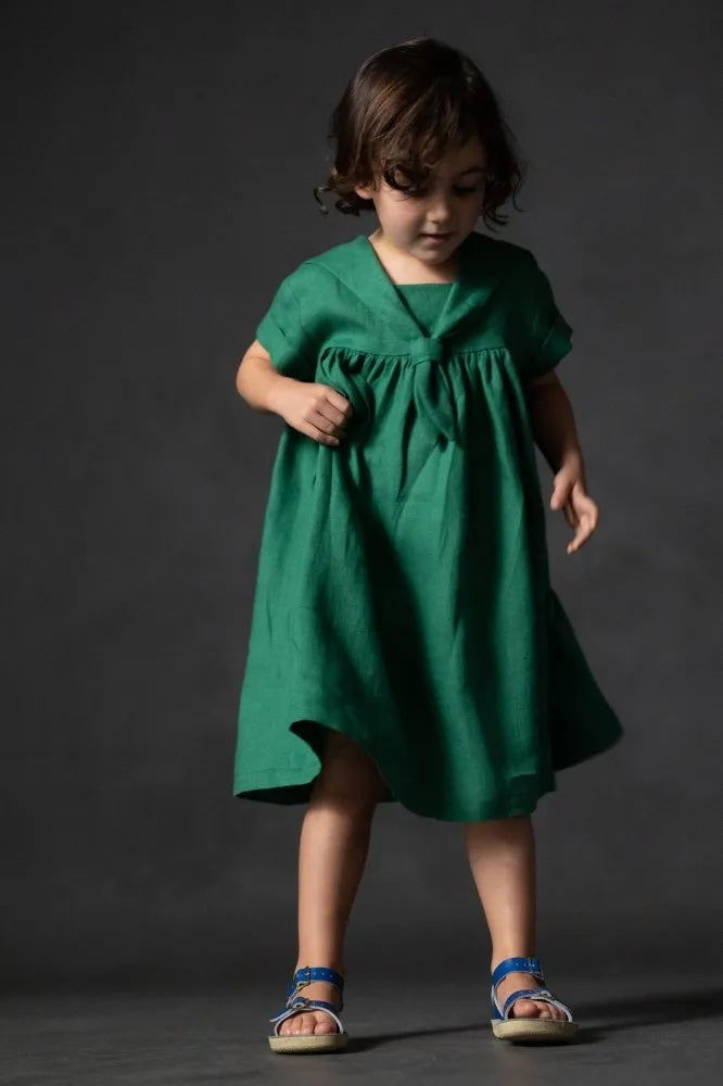 Girl wearing the Children's Skipper Dress sewing pattern from Merchant & Mills on The Fold Line. A dress pattern made in denim, cotton chambray, laundered linen, or lightweight wool fabric, featuring a relaxed fit, dropped shoulders, short sleeves with rolled cuffs, and a sailor collar.