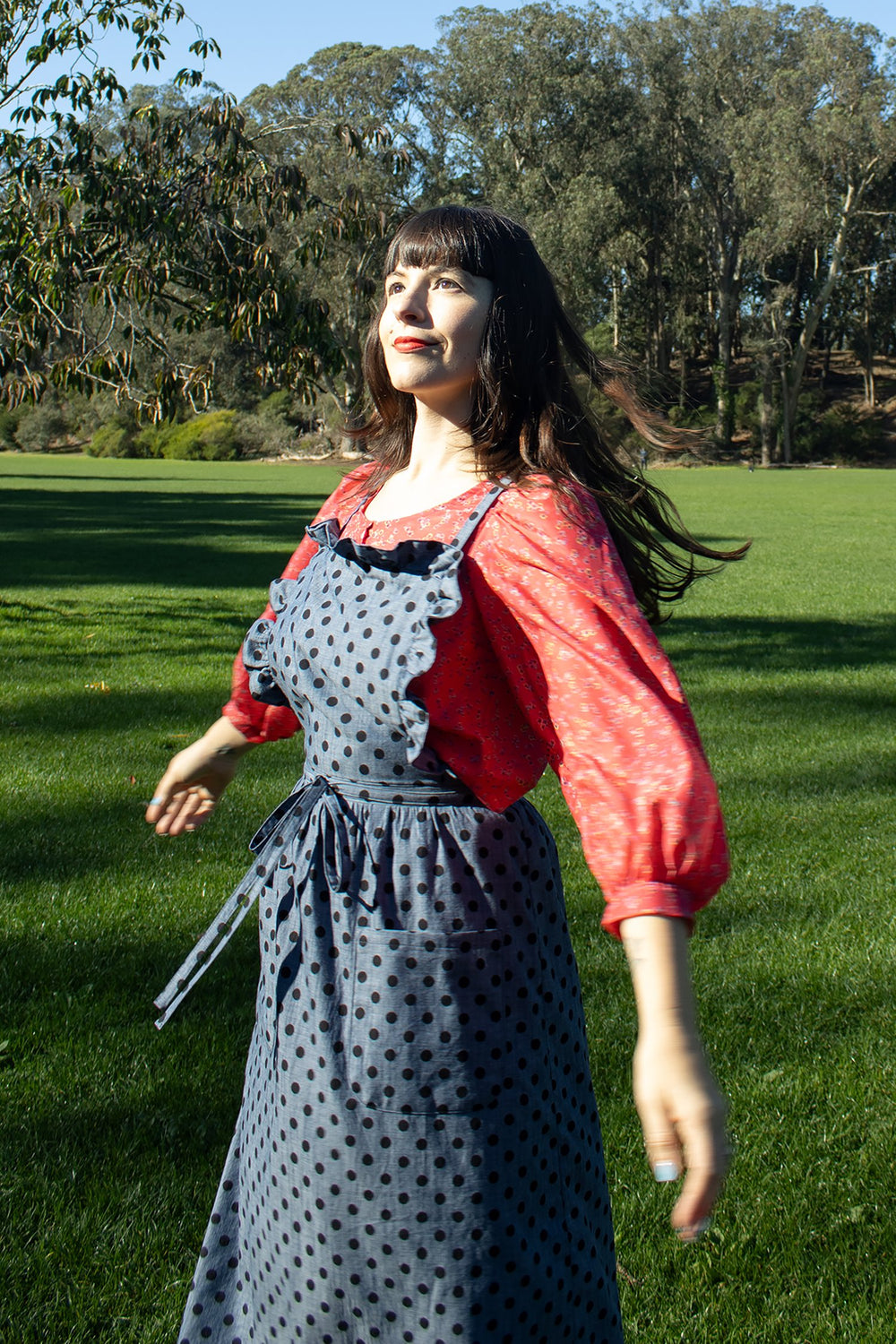 Woman wearing the Dillon Dress sewing pattern from Christine Haynes on The Fold Line. A pinafore or apron dress pattern made in linen, cotton, denim, or corduroy fabric, featuring a bib with a ruffle, thin shoulder straps, a gathered A-line skirt, skinny waistband that ties in front, and patch pockets.