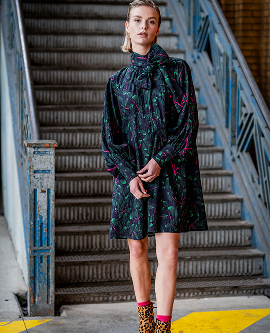 Woman wearing the Electra Dress sewing pattern from Fibre Mood on The Fold Line. A dress pattern made in seersucker, double gauze, crepe, broderie anglaise, poplin, or woven jacquard fabric, featuring a voluminous silhouette, statement pussy bow, wide sleeves with fitted cuffs, and above knee length.