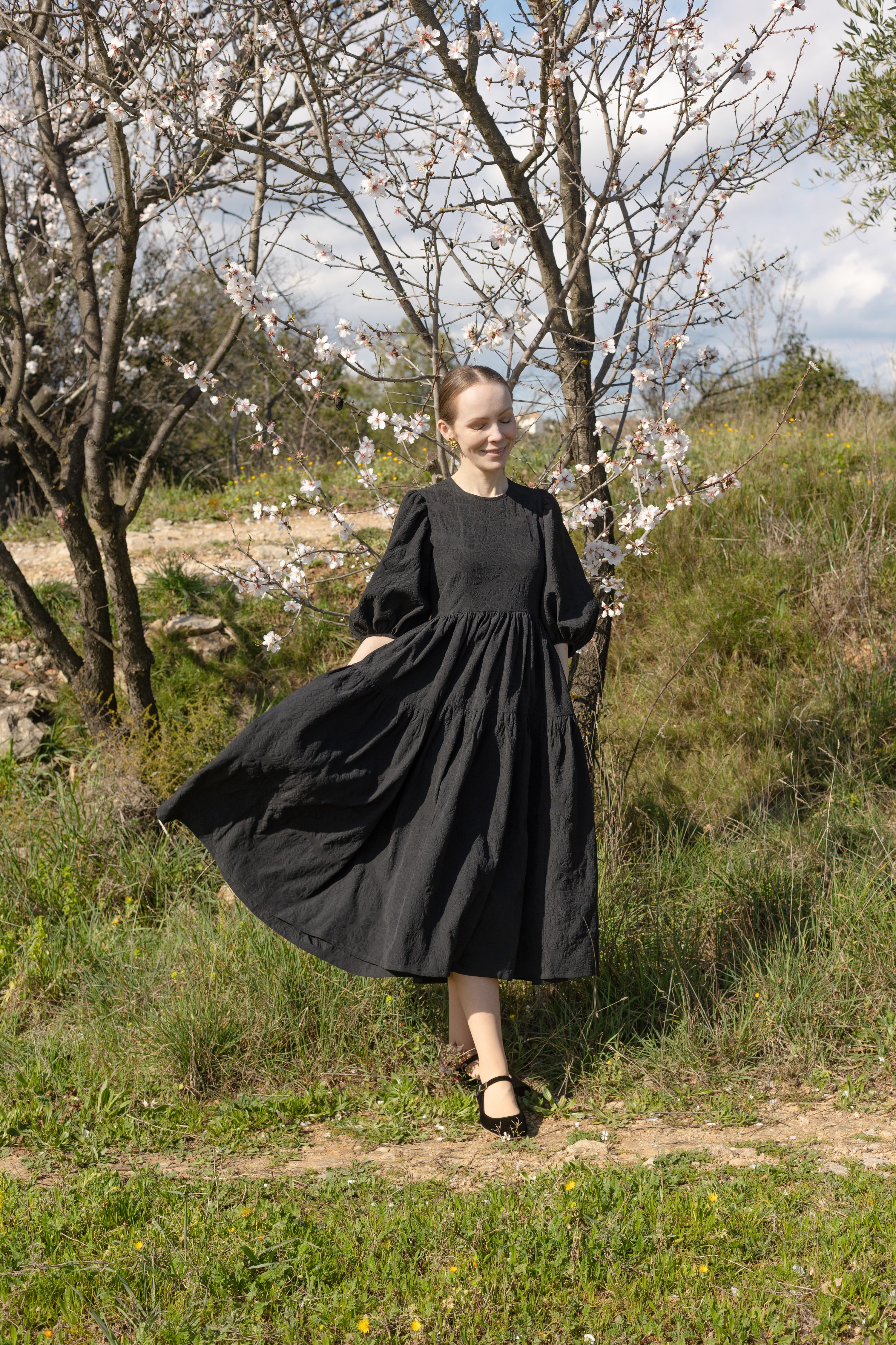 Woman wearing the Elsa Dress sewing pattern from Silversaga Patterns on The Fold Line. A dress pattern made in cotton, linen, or wool fabric, featuring a round neckline, mid-length statement sleeves with a dipped hemline, high curved waistline, and a tiered midi length gathered skirt.