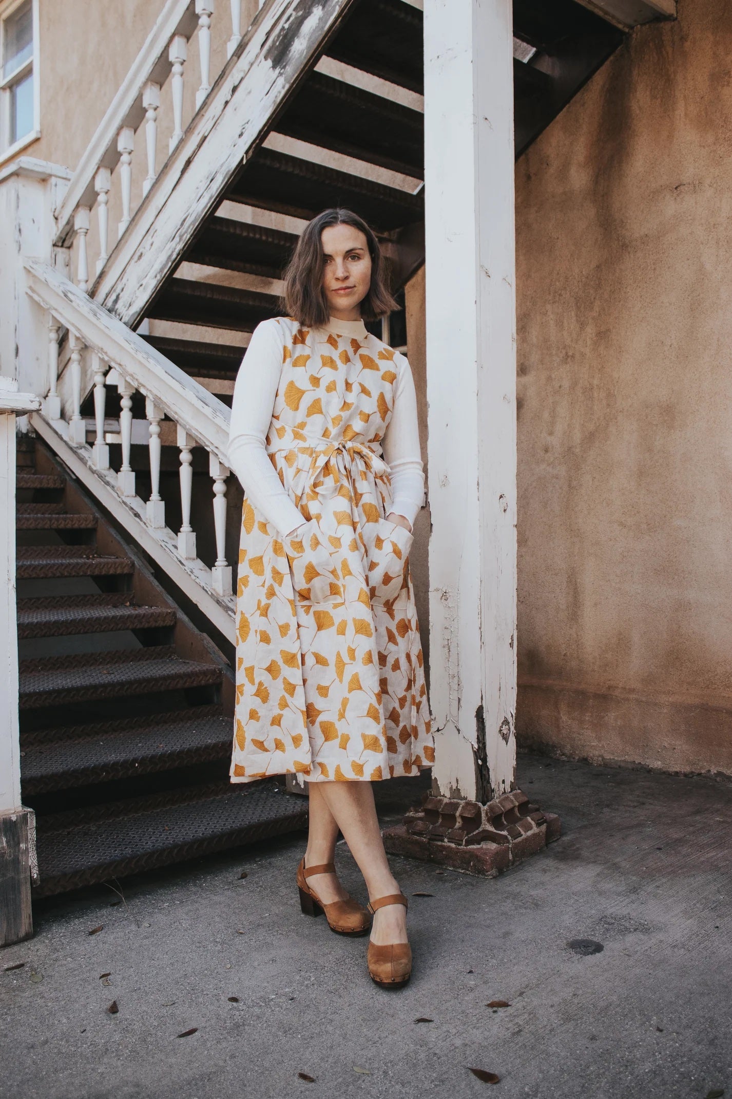 Woman wearing the Ginkgo Pinafore sewing pattern from Madswick on The Fold Line. A sleeveless wrap dress pattern made in linen, cotton, tencel, viscose, viscose blend, wool, or denim fabric, featuring a fully lined dartless bodice, square neckline, large patch pockets, a gathered skirt, adjustable ties, and midi length.