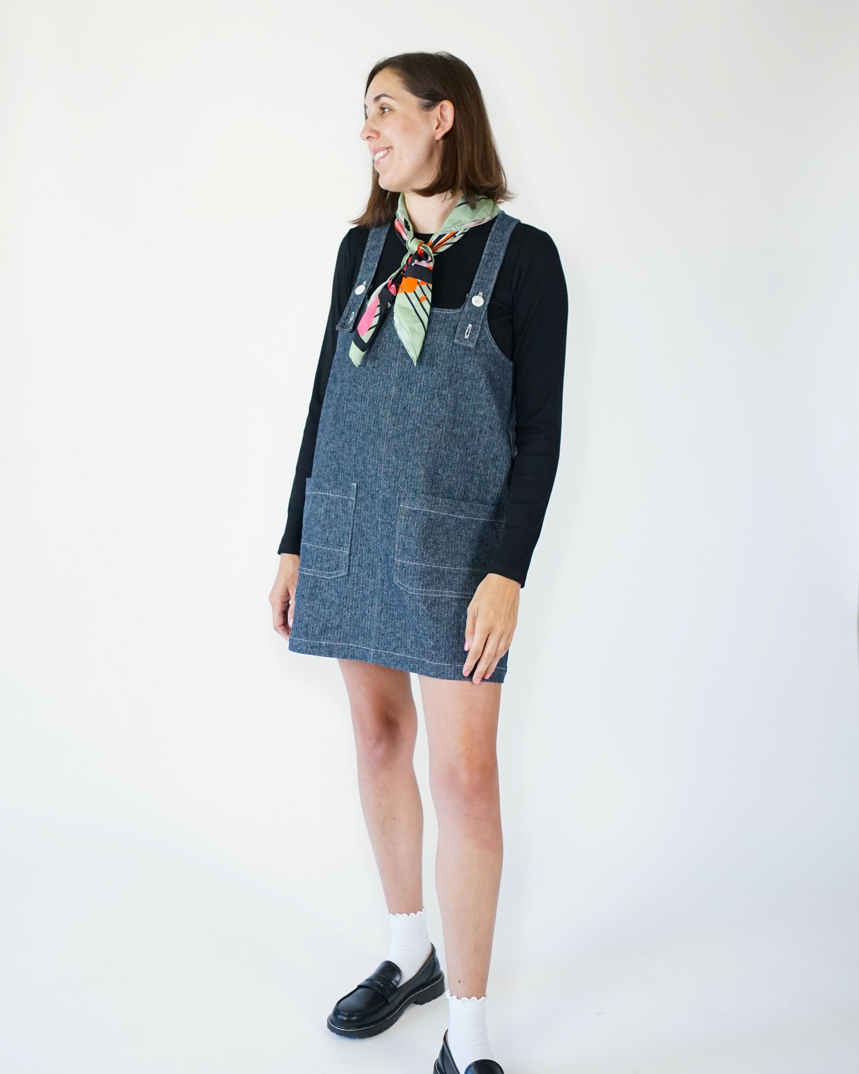 Woman wearing the Hopscotch Jumper sewing pattern from Matchy Matchy Sewing Club on The Fold Line. A pinafore dress pattern made in twill, denim, or canvas fabric, featuring a subtle A-line shape, adjustable straps, patch pockets, and mini length.