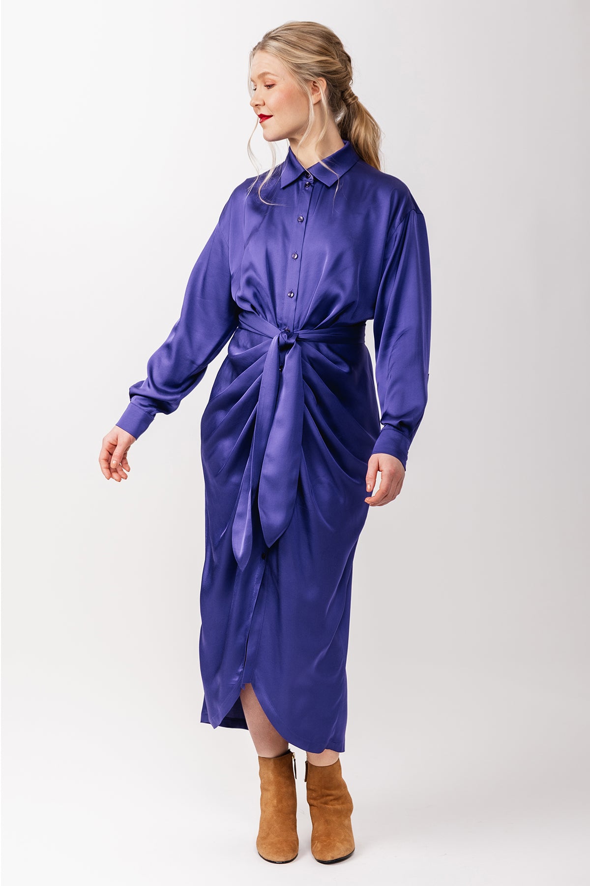 Woman wearing the Ilta Shirt Dress sewing pattern from Named on The Fold Line. A dress pattern made in satin, viscose challis, or crepe fabric, featuring a collar, dropped shoulders, pleated front for a ruched effect, button fastening, long sleeves with cuffs, rounded front hem, and midi length.