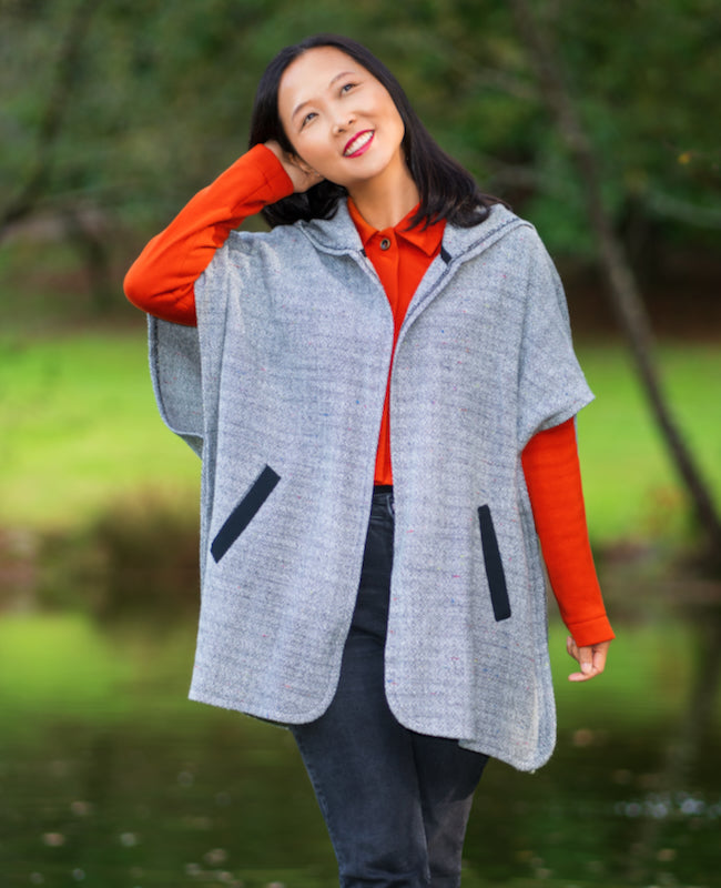 Woman wearing the Koersel Cape sewing pattern from Itch to Stitch on The Fold Line. A cape pattern made in coating, wool blend, or double knit fabric, featuring an open front, elbow-length sleeves, an oversized hood, welt pockets, and mid-thigh length.