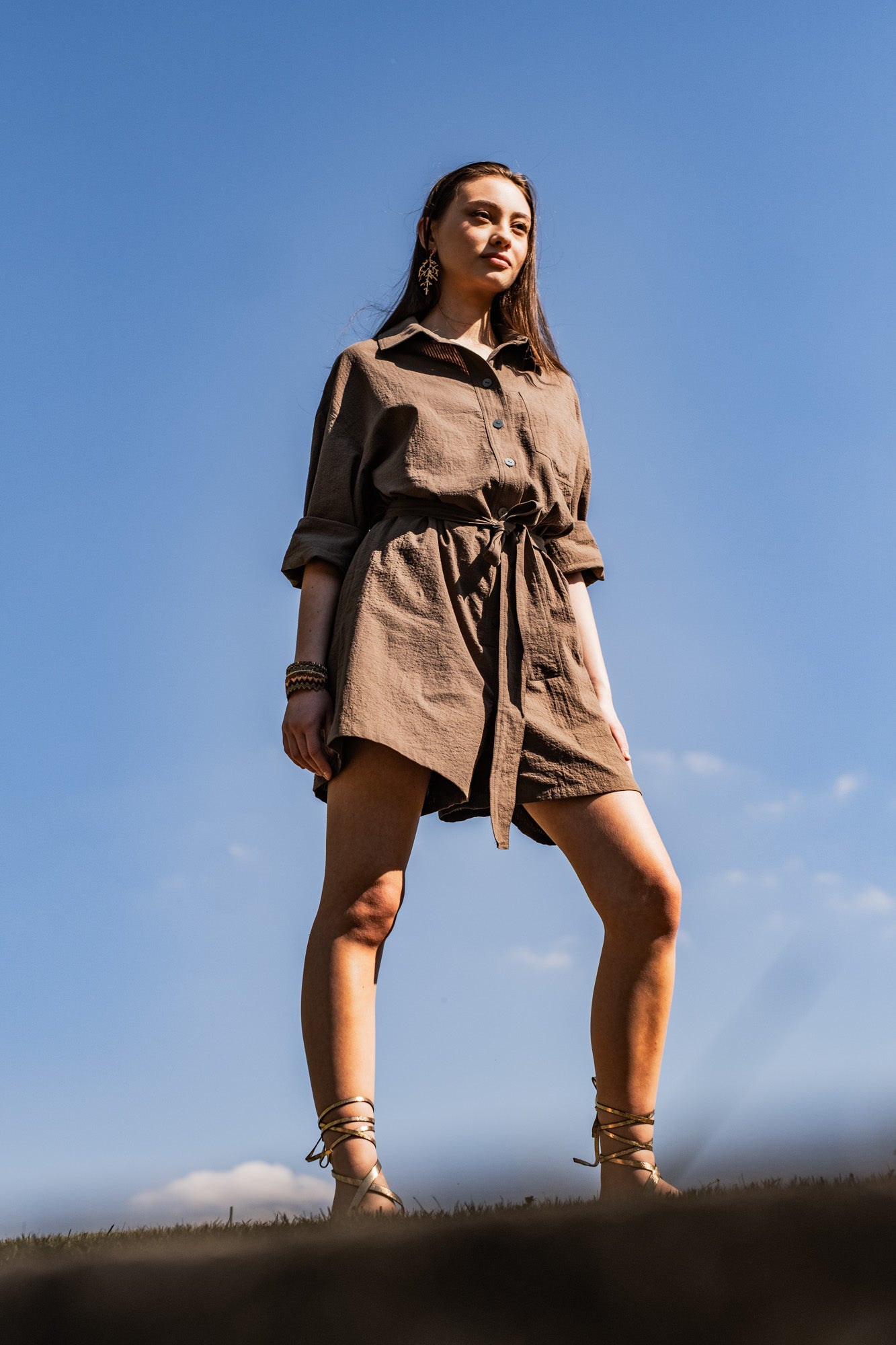 Woman wearing the Liberty Playsuit sewing pattern from Fibre Mood on The Fold Line. A playsuit pattern made in cotton twill, poplin, fine corduroy, or linen fabric, featuring an oversized fit, shirt collar, front button placket, long sleeves with cuffs, chest pocket, tie belt, and shorts length.