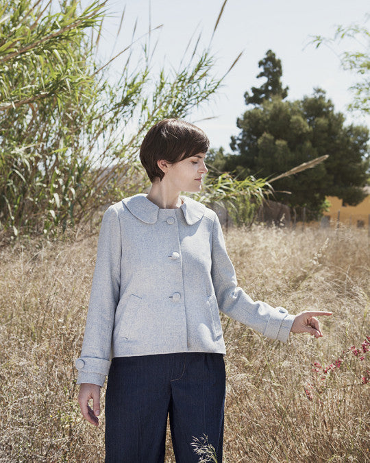 Woman wearing the Ninot Jacket sewing pattern from Pauline Alice on The Fold Line. A jacket pattern made in woollen, felt, boiled wool, tweed, or gabardine fabric, featuring a round collar, shoulder yoke, long sleeves, button tabs, and welt pockets.