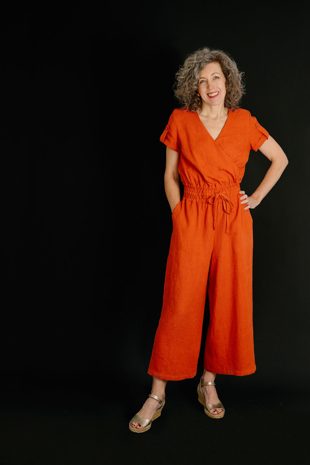 Woman wearing the Northbridge Jumpsuit sewing pattern from Liesl + Co on The Fold Line. A jumpsuit pattern made in linen, cotton twill, poplin, shirting, or rayon fabric, featuring a mock-wrap bodice, cuffed short sleeves with button tabs, smocked waist with drawstring ties, and cropped wide legs.