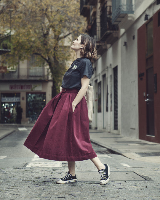 Woman wearing the Riola Skirt sewing pattern from Pauline Alice on The Fold Line. A skirt pattern made in poplin, twill, linen, chambray, or light woollen fabric, featuring a high waist, gathers, front inverted pleat, hidden front opening, shaped buttoned waistband, in-seam pockets, and midi length.