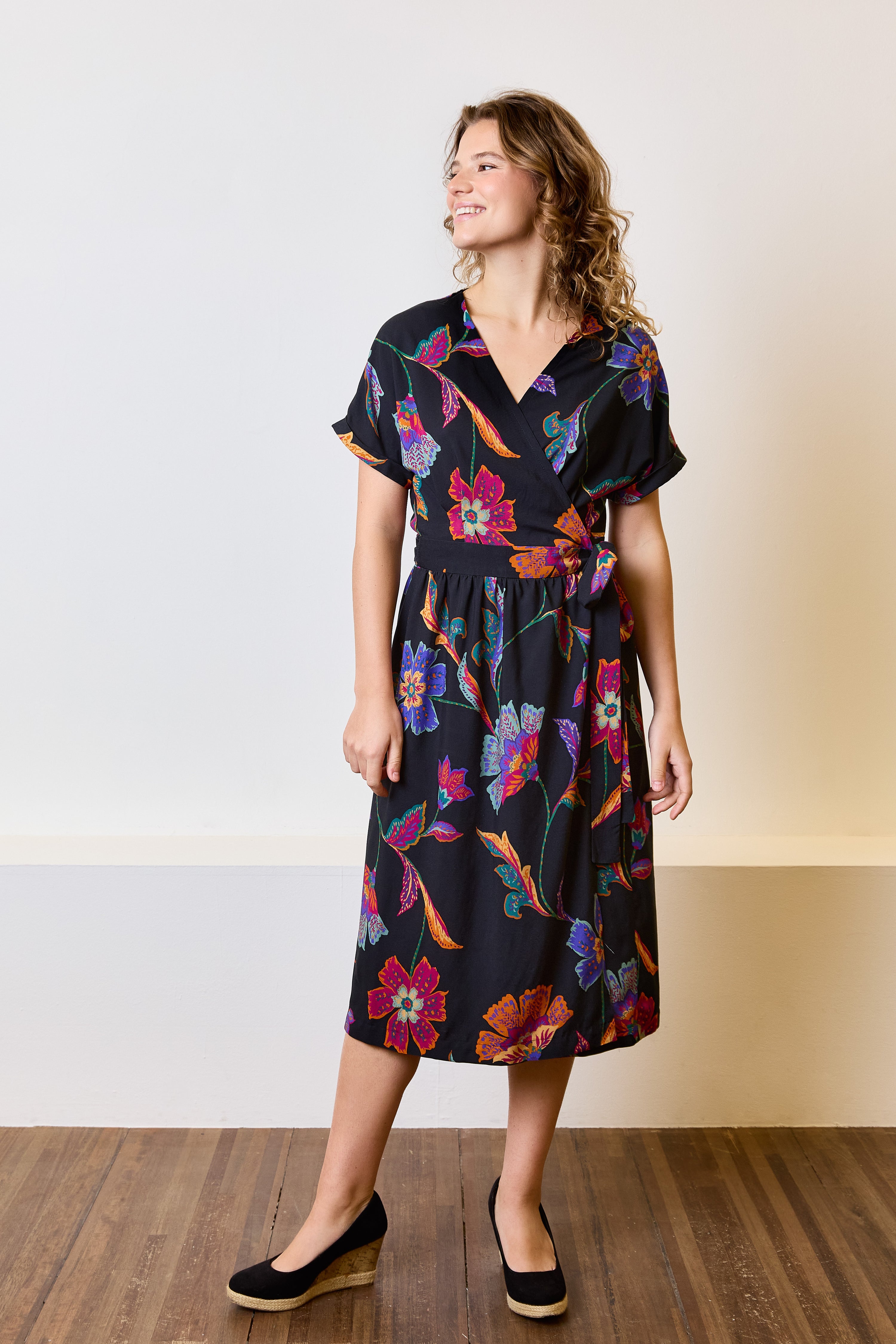 Woman wearing the Robyn Wrap Dress sewing pattern from Atelier Jupe on The Fold Line. A dress pattern made in viscose, cotton, linen, tencel, or polyester fabric, featuring a V-neck, short grown-on sleeves with cuffs, back darts, waist ties, and a gathered skirt with a large overlap.
