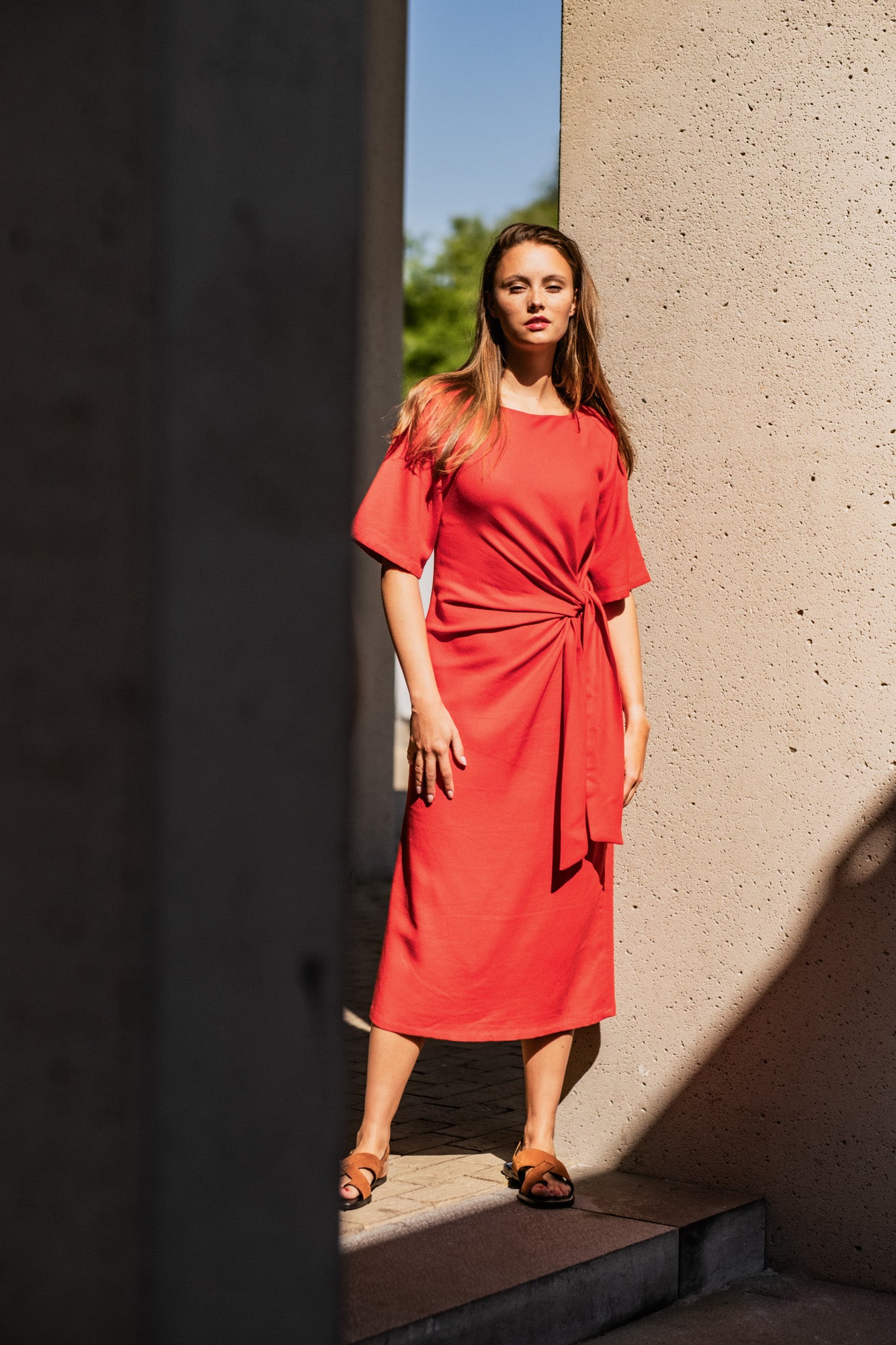 Woman wearing the Suvi Dress sewing pattern from Fibre Mood on The Fold Line. A dress pattern made in crepe, lyocell blend, cupro, satin, linen, viscose twill, or medium weight knit fabric, featuring a round neckline, asymmetrical ties at the waist, mid-length sleeves, back slit, and midi length.