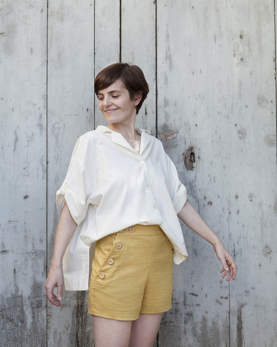 Woman wearing the Vera Shirt sewing pattern from Pauline Alice on The Fold Line. A shirt pattern made in cotton lawn, linen, rayon, tencel, cupro, crepe, or poplin fabric, featuring a loose fit, notched collar, soft gathers at the back neckline, short sleeves with button tabs, and a high-low hem.