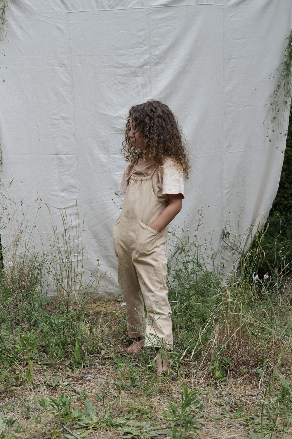 Child wearing the Children's Patsy Overalls sewing pattern from Ready to Sew on The Fold Line. An overall/dungaree pattern made in cotton poplin, linen, batik, light denim or corduroy fabrics, featuring a relaxed fit, full length leg, front slash pockets,