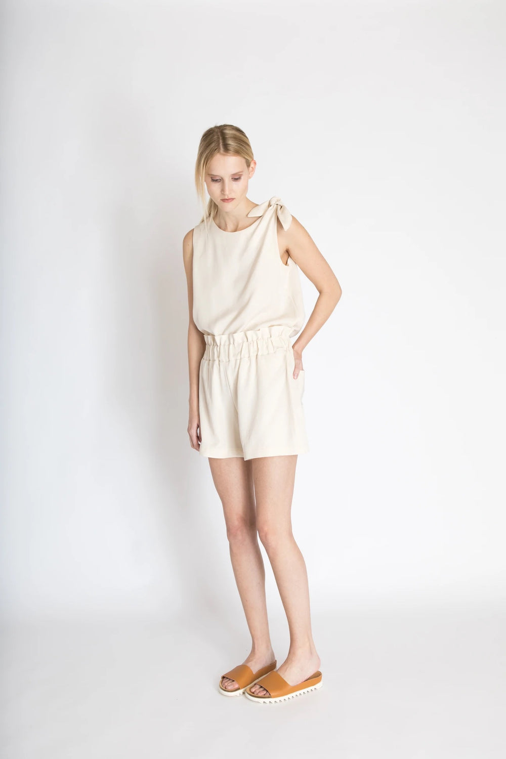 Woman wearing the Freja Knot Top sewing pattern from Bara Studio on The Fold Line. A sleeveless top pattern made in cotton, linen or tencel fabrics, featuring a loose-fit, knot on the left shoulder and scoop neckline.