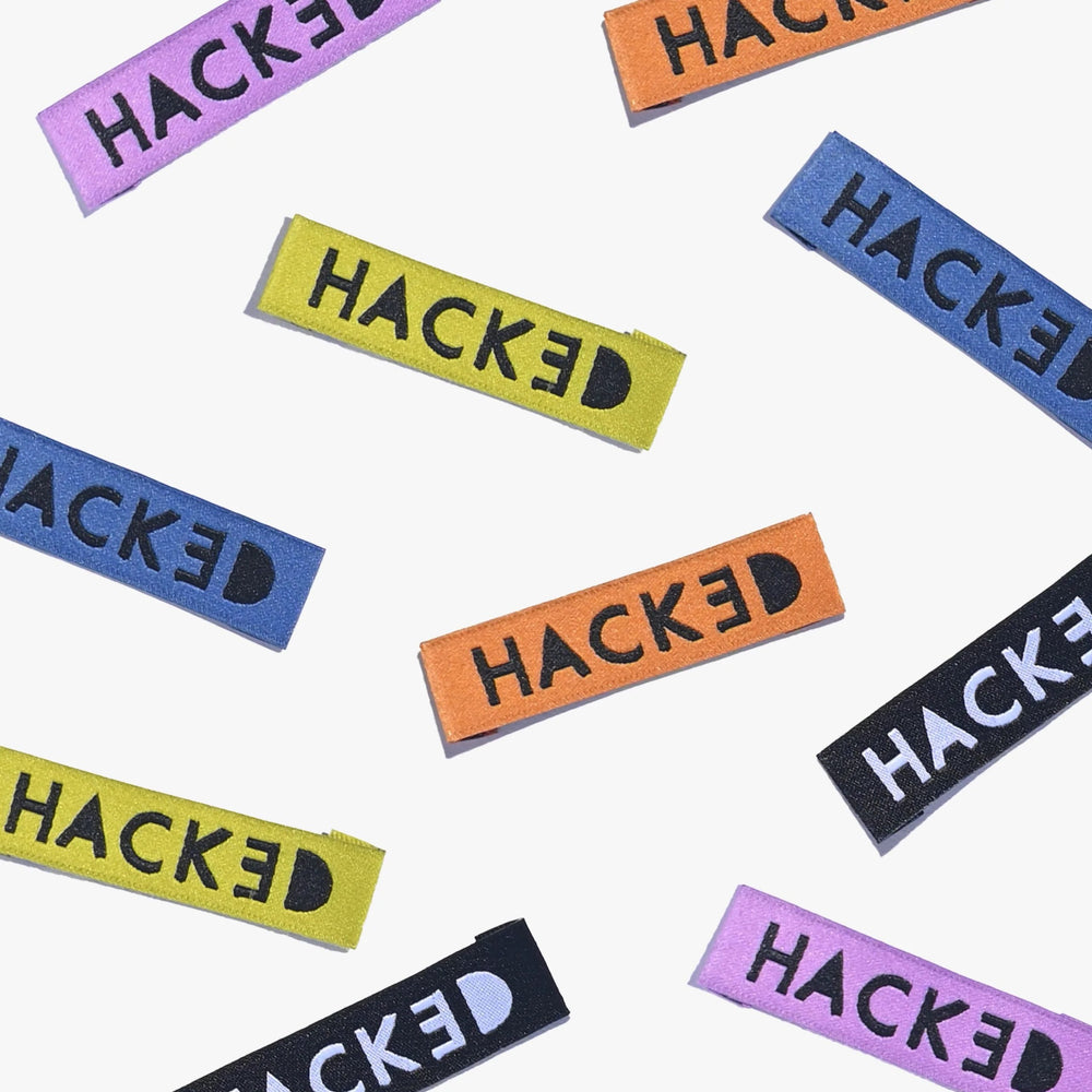 Photo showing the 'Hacked' Cotton Labels from Kylie & The Machine on The Fold Line. A washable, durable, and non-scratchy cotton label featuring 10 labels with black, blue, orange, purple or chartreuse coloured backgrounds, they are all ready to be sewn i