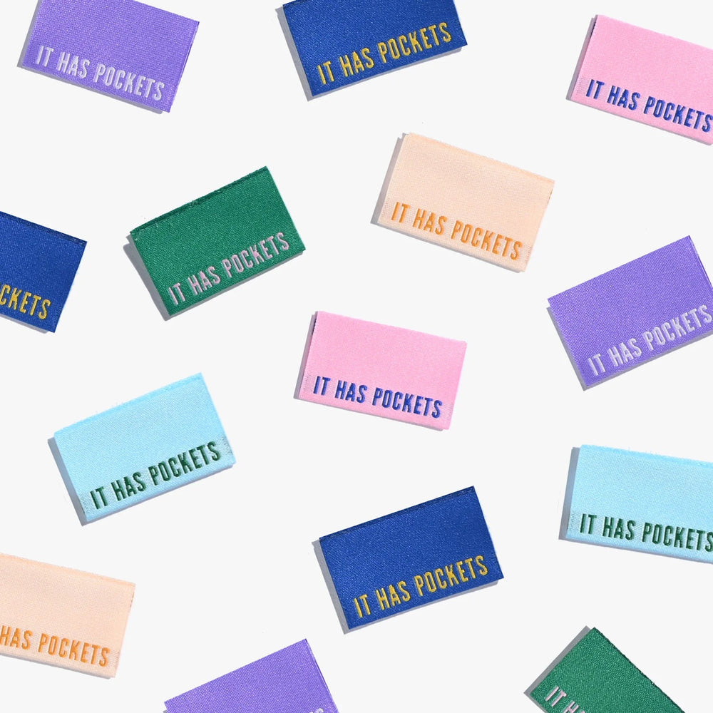Photo showing 'It Has Pockets' Cotton Labels from Kylie & The Machine on The Fold Line. A washable, durable, and non-scratchy cotton label featuring multi-coloured backgrounds and multi-coloured text, they are all ready to be sewn into your handmade cloth