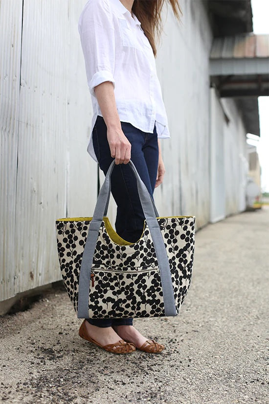 Woman holding the Poolside Tote sewing pattern from Noodlehead on The Fold Line. A tote pattern made in heavy linen, denim, canvas, twill or duck cloth fabrics, featuring a roomy size, inside key loop, exterior pocket with zip closure and shoulder length