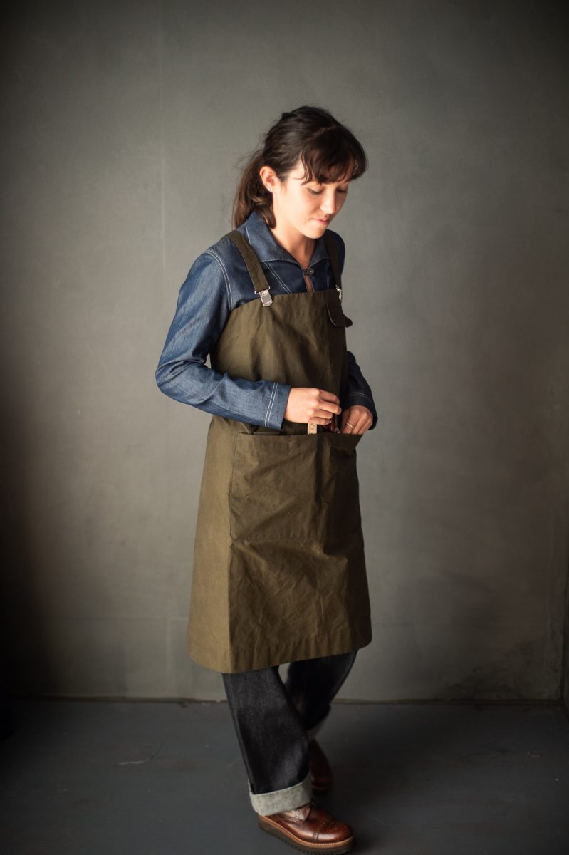 Woman wearing the Victor Apron sewing pattern by Merchant and Mills. An apron pattern made in oilskin, denim, cotton canvas or drill fabric featuring a large divided waist pocket with scissor holder, top pocket and cross back for comfort.