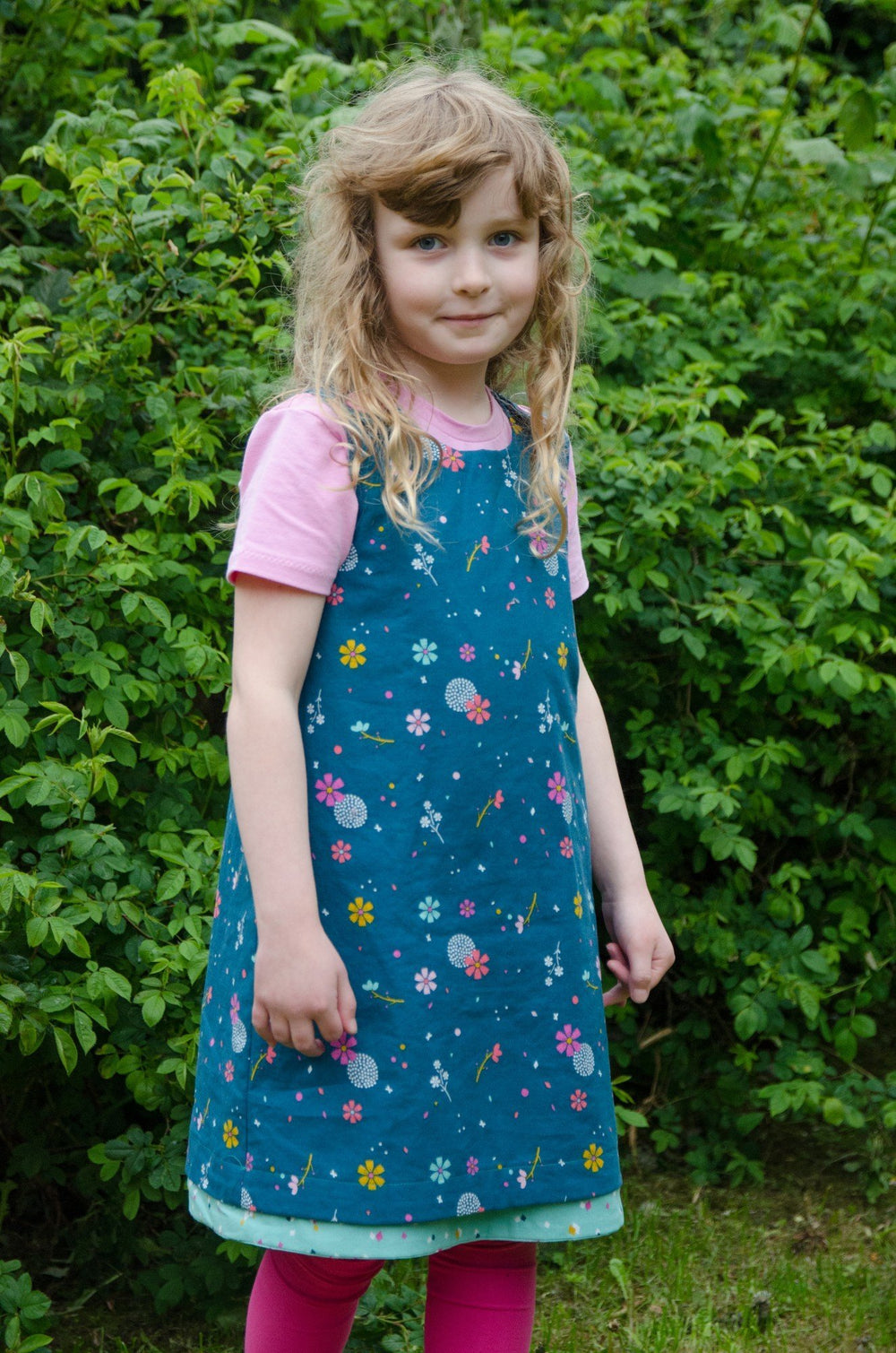 Child wearing the Baby/Child Rosie Dress sewing pattern from Bobbins and Buttons on The Fold Line. A pinafore dress pattern made in cottons, lightweight denim, broadcloth, corduroy, needlecord or denim fabrics, featuring button through straps, round neck