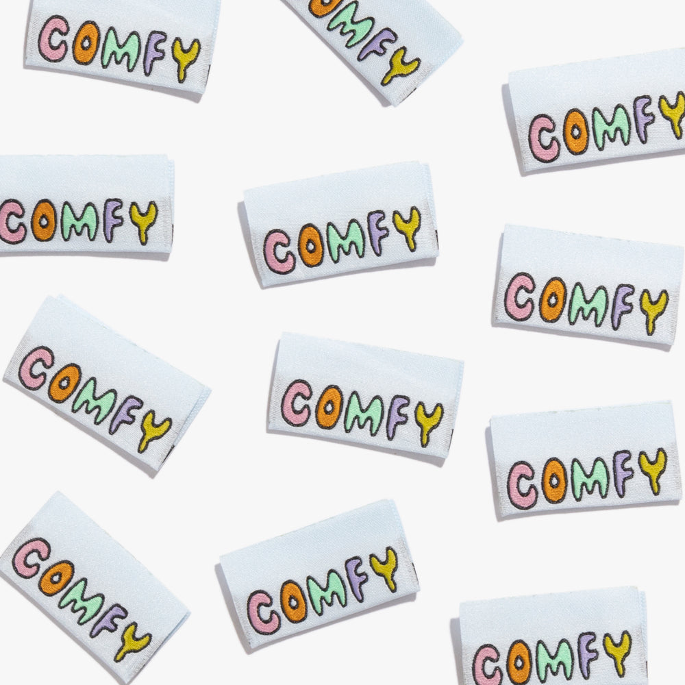 Photo showing 'Comfy' Woven Labels from Kylie & The Machine on The Fold Line. A washable, durable and non-scratchy fabric label featuring a baby blue background, and the word ‘Comfy’ in multi-coloured text all ready to be sewn into your handmade clothes.