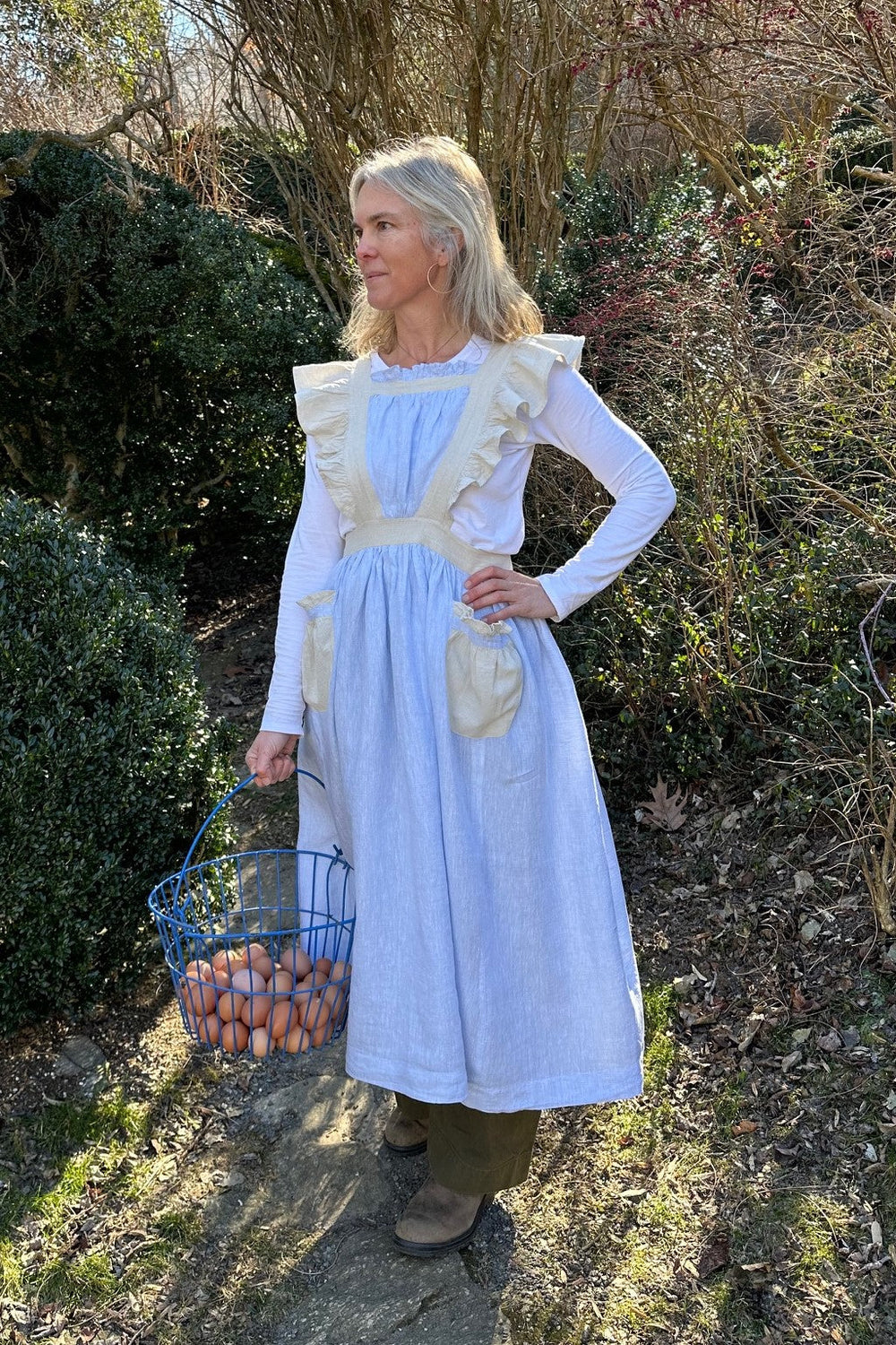 Woman wearing the 303 English Cottage Kitchen Apron sewing pattern from Folkwear on The Fold Line. An apron pattern made in cottons, linen, silks or rayon fabrics, featuring a midi length ruffled pinafore apron with pockets, plus biscuit cozy, napkins, te