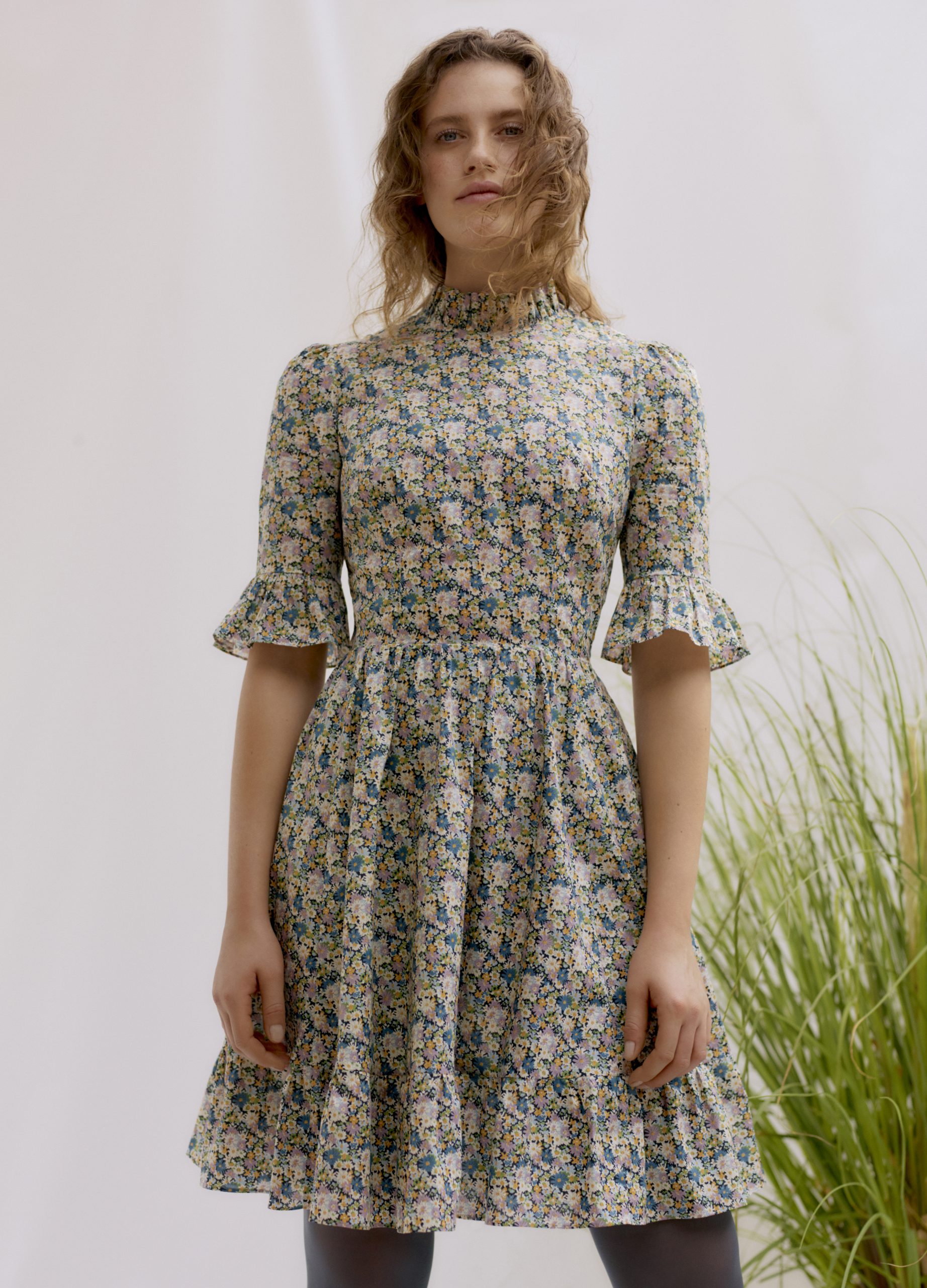 Woman wearing the Alexa Frill Dress sewing pattern by Liberty Sewing Patterns. A dress pattern made in cotton, satin or poplin fabrics, featuring elbow length sleeves with frill, stand frill collar, fitted bodice, flared skirt with tiered frill and back z