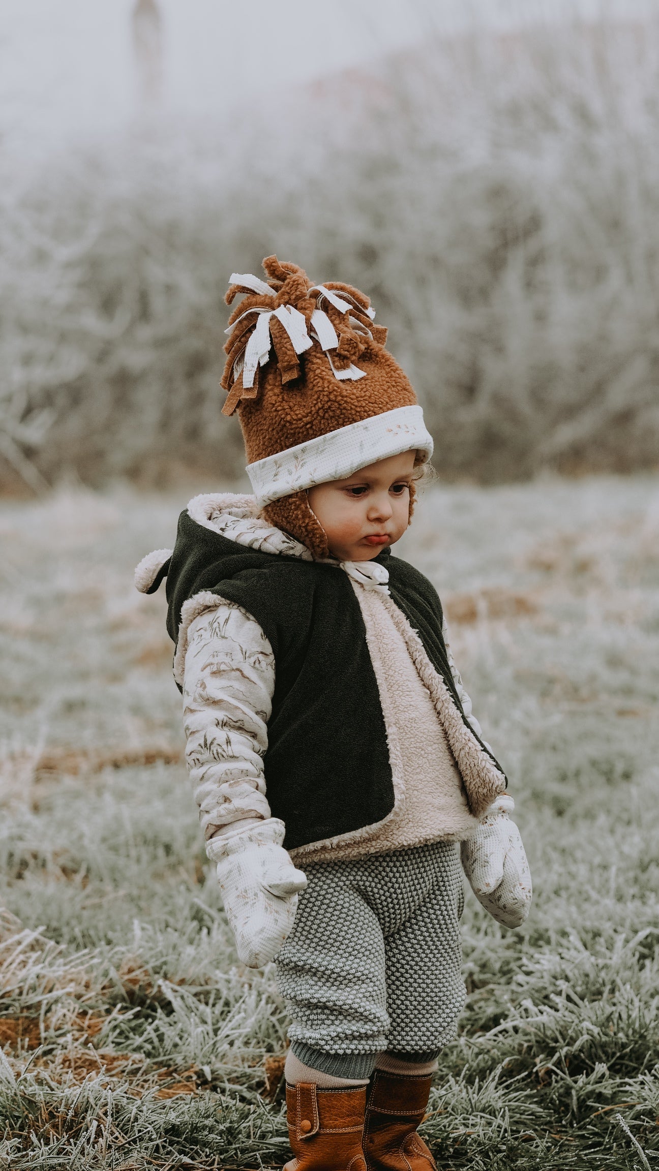 Child wearing the Baby/Child Bidibou Hat and Mittens sewing pattern from Petits D’om on The Fold Line. A hat and mittens pattern made in minkee, jersey, sweat fabric, velvet or stretch fleece fabrics, featuring a hat with ear flaps and fabric pompom on to