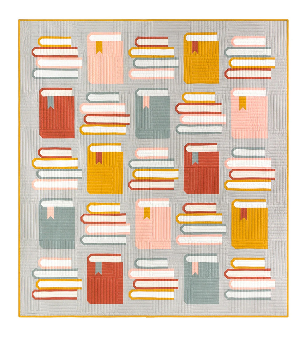 Photo showing the Book Nook Quilt sewing pattern from Pen and Paper Patterns on The Fold Line. A quilt pattern made in quilting cotton fabrics, featuring an alternating upright book with a four stack of books in blues, reds, yellows and pinks on a grey ba