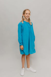 Girl wearing the 110 Children's Little Kittel sewing pattern from Folkwear on The Fold Line. A dress pattern made in linen, homespun, broadcloth, gingham, denim, featherweight corduroy, or lightweight wool fabric, featuring a neckband with pleats, long sleeves with buttoned cuffs, and knee length.
