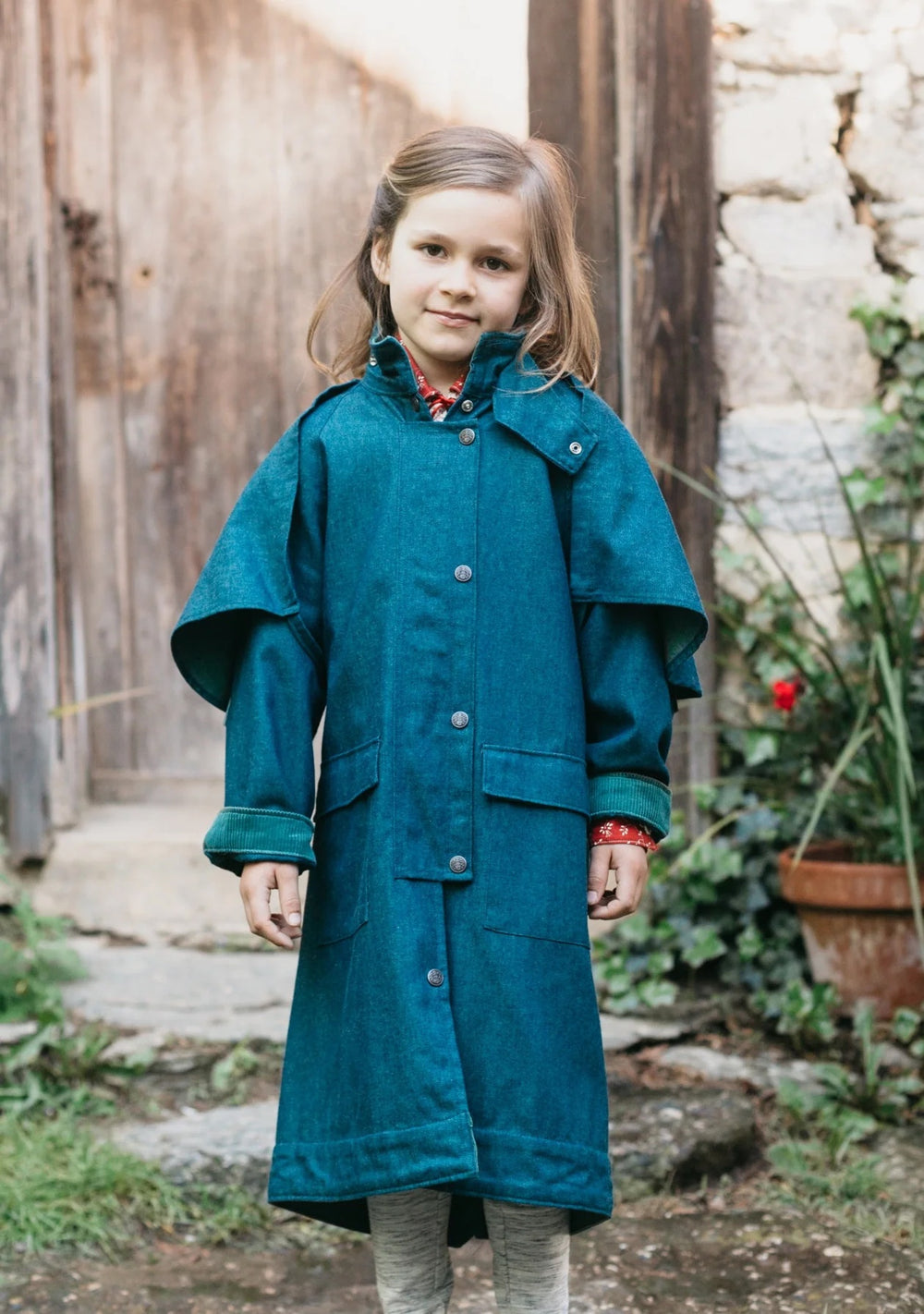 Girl wearing the 138 Child/Teen Australian Drover's Coat sewing pattern from Folkwear on The Fold Line. A coat pattern made in denim, canvas, poplin, wool, linen, ripstop, or other waterproof fabric, featuring a high neck, raglan sleeves, detachable shoulder cape, snap front, pockets, and partial lining.
