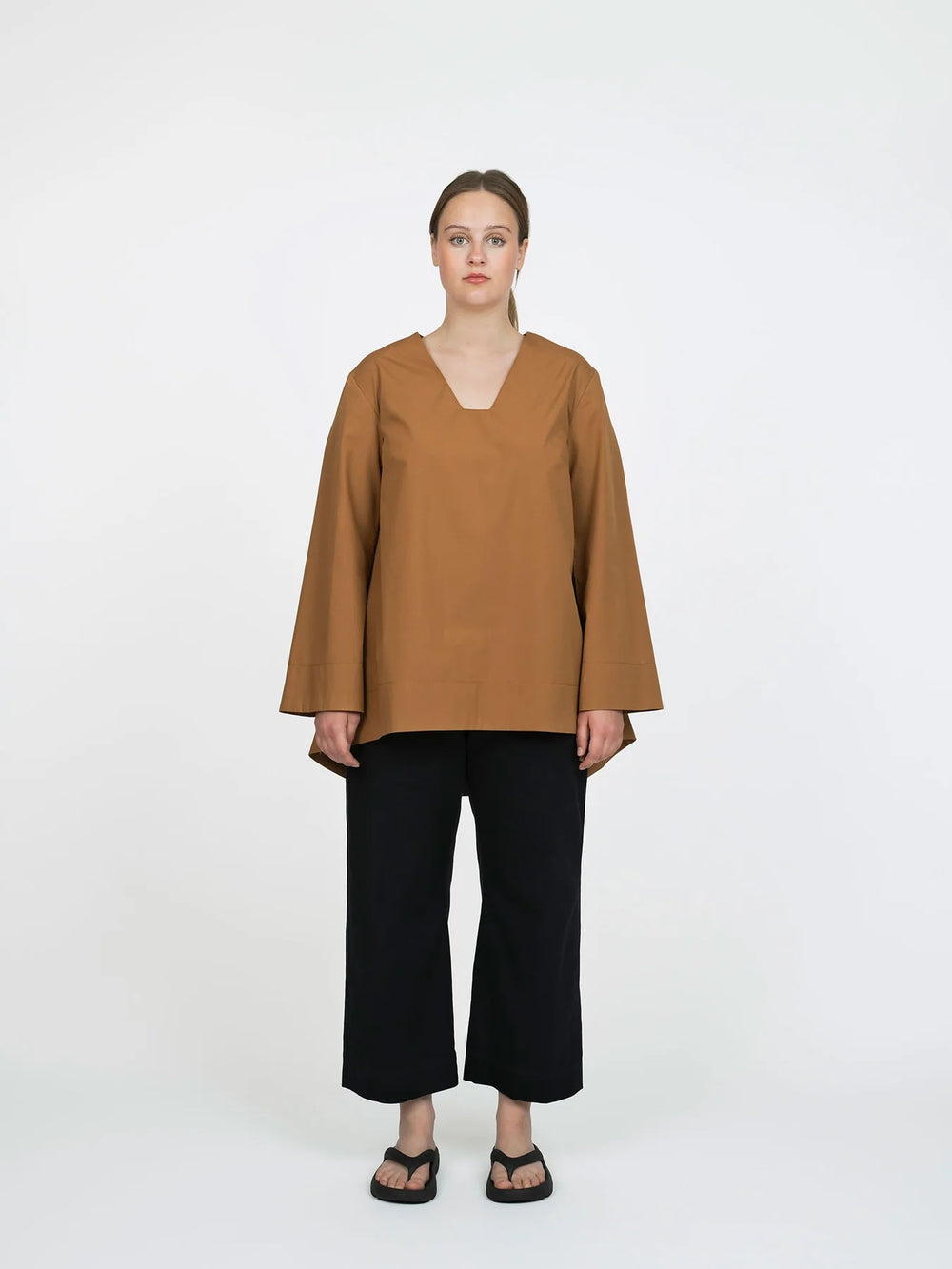Woman wearing the A-line Tunic sewing pattern from The Assembly Line on The Fold Line. A top pattern made in light to medium weight fabric, featuring a relaxed fit, V-shaped neckline with a square-cut bottom, dropped shoulders, long sleeves, and a dipped back hem.