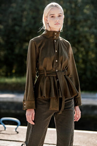 Woman wearing the Aki Shirt sewing pattern from Fibre Mood on The Fold Line. A shirt pattern made in leather, suede, or denim fabric, featuring a high neck, button front, long sleeves with cuffs, matching tie belt, and peplum.