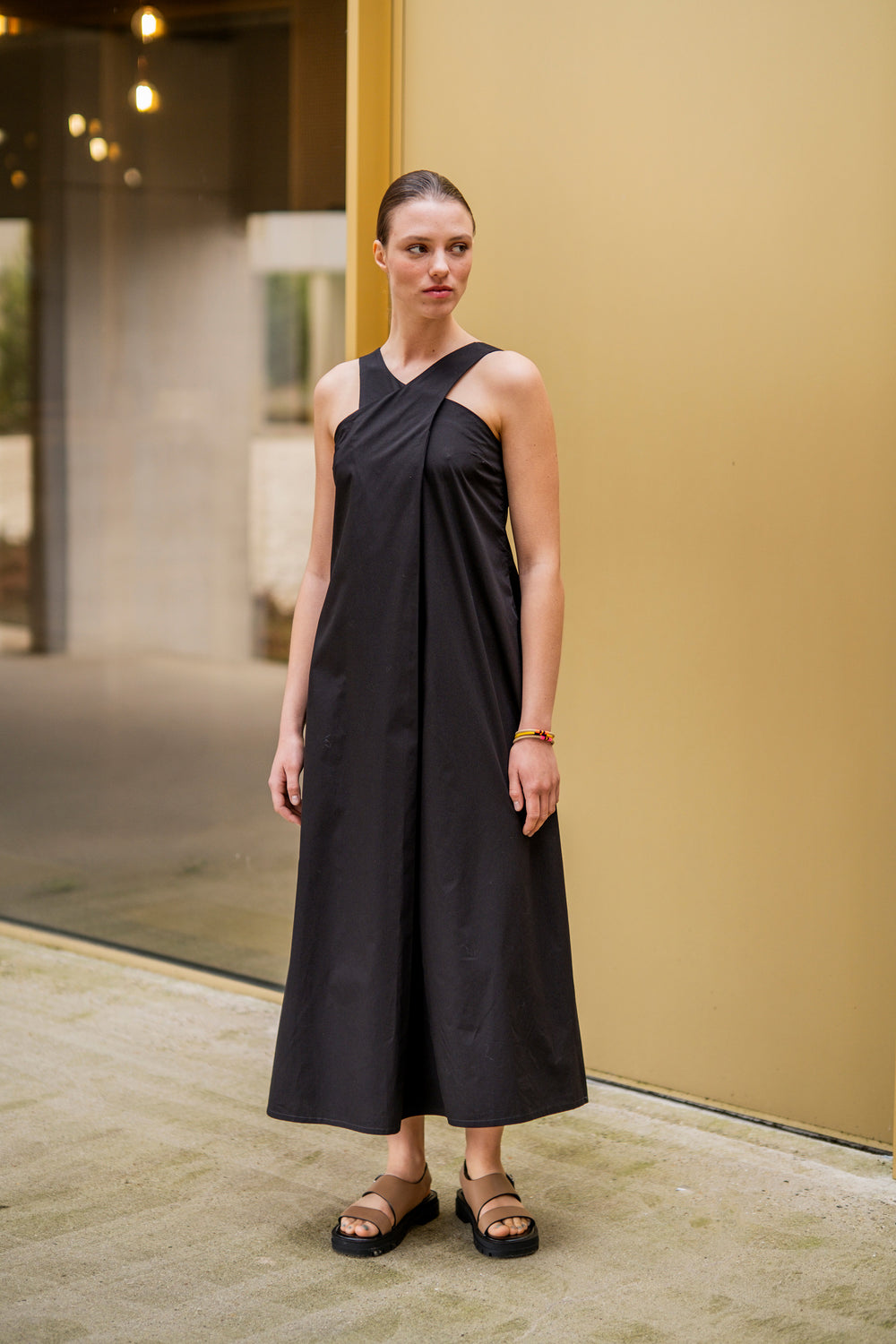 Woman wearing the Alberthe Dress sewing pattern from Fibre Mood on The Fold Line. A sleeveless dress pattern made in poplin, cotton twill, polycotton, denim, linen, double gauze, seersucker, chambray, lyocell, or crepe fabric, featuring a crossed neckline at the front, concealed zipper in the side seam, and midi length.