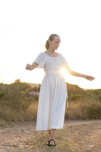 Woman wearing the Alma Dress sewing pattern from Silversaga Patterns on The Fold Line. A dress pattern made in linen, cotton, block prints, rayon/viscose, or tencel fabric, featuring a gathered round neckline, flutter sleeves, an adjustable drawstring waist with fabric ties at the sides, an A-line skirt, and in-seam pockets.