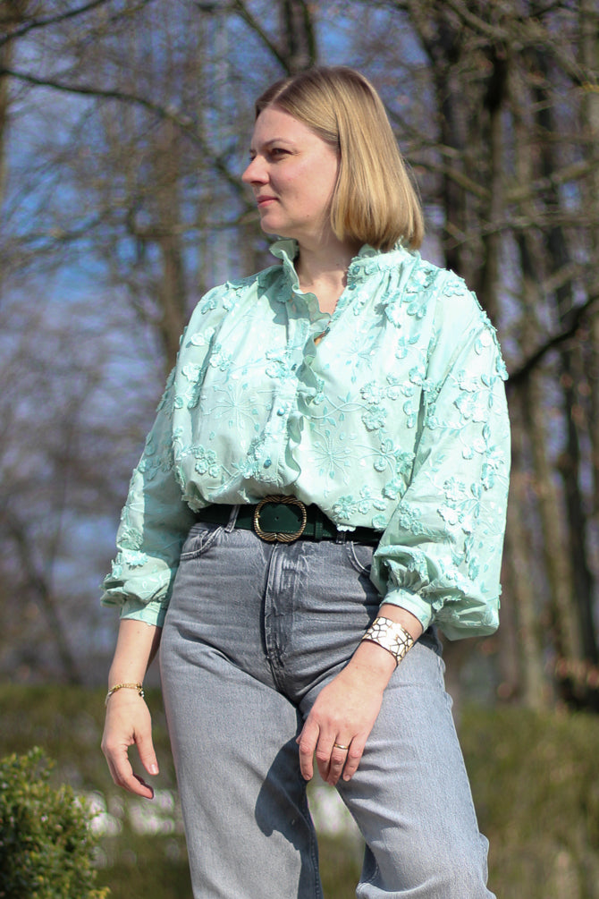 Woman wearing the Anoushka Blouse sewing pattern from Lenaline Patterns on The Fold Line. A shirt pattern made in cotton poplin, cotton twill, linen, tencel, denim, cotton twill, chino, flannel, viscose, or silk fabric, featuring a relaxed fit, high collar, gathers at the front and back, a gathered ruffle, button placket, and long sleeves with cuffs.