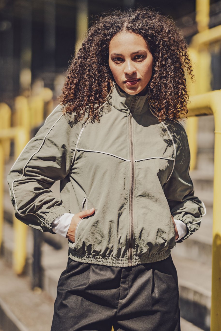 Woman wearing the Ash Jacket sewing pattern from Fibre Mood on The Fold Line. A jacket pattern made in polyester microfibre, nylon, softshell, French terry, heavy jersey, or sweater fabric, featuring a relaxed fit, stand-up collar, front zip, raglan sleeves, zipped pockets, elasticated hems, and piping along the seams.