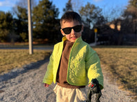 Boy wearing the Baby/Child Mini Cardamom Quilt Jacket sewing pattern from Sew Bake Make on The Fold Line. A reversible jacket pattern made in a lightweight thrifted quilt or quilted fabric, featuring a roomy fit, stand collar, and pockets.
