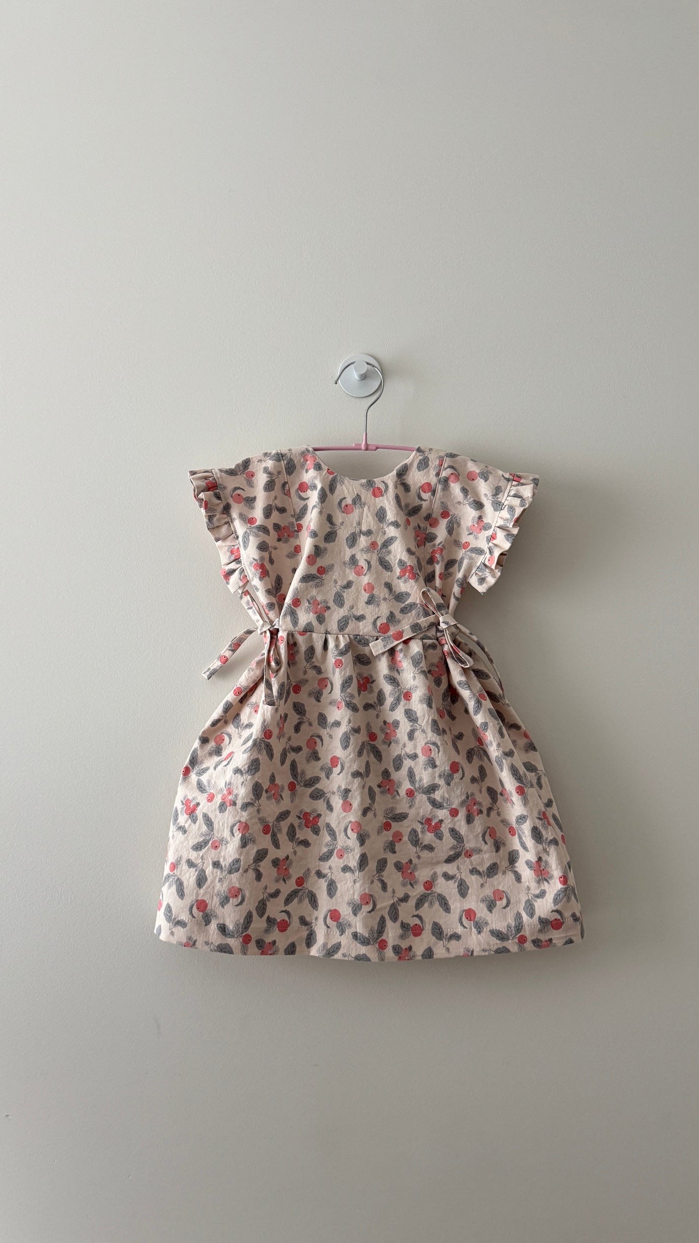 The Baby/Child Mini Tie Dress sewing pattern from Sewing Therapy on The Fold Line. A dress pattern made in cotton, double gauze, cotton-linen blend, muslin, cotton jersey, voile, corduroy, or flannel fabric, featuring a reversible neckline, button closure, ruffled sleeves, gathered skirt, and waist ties.