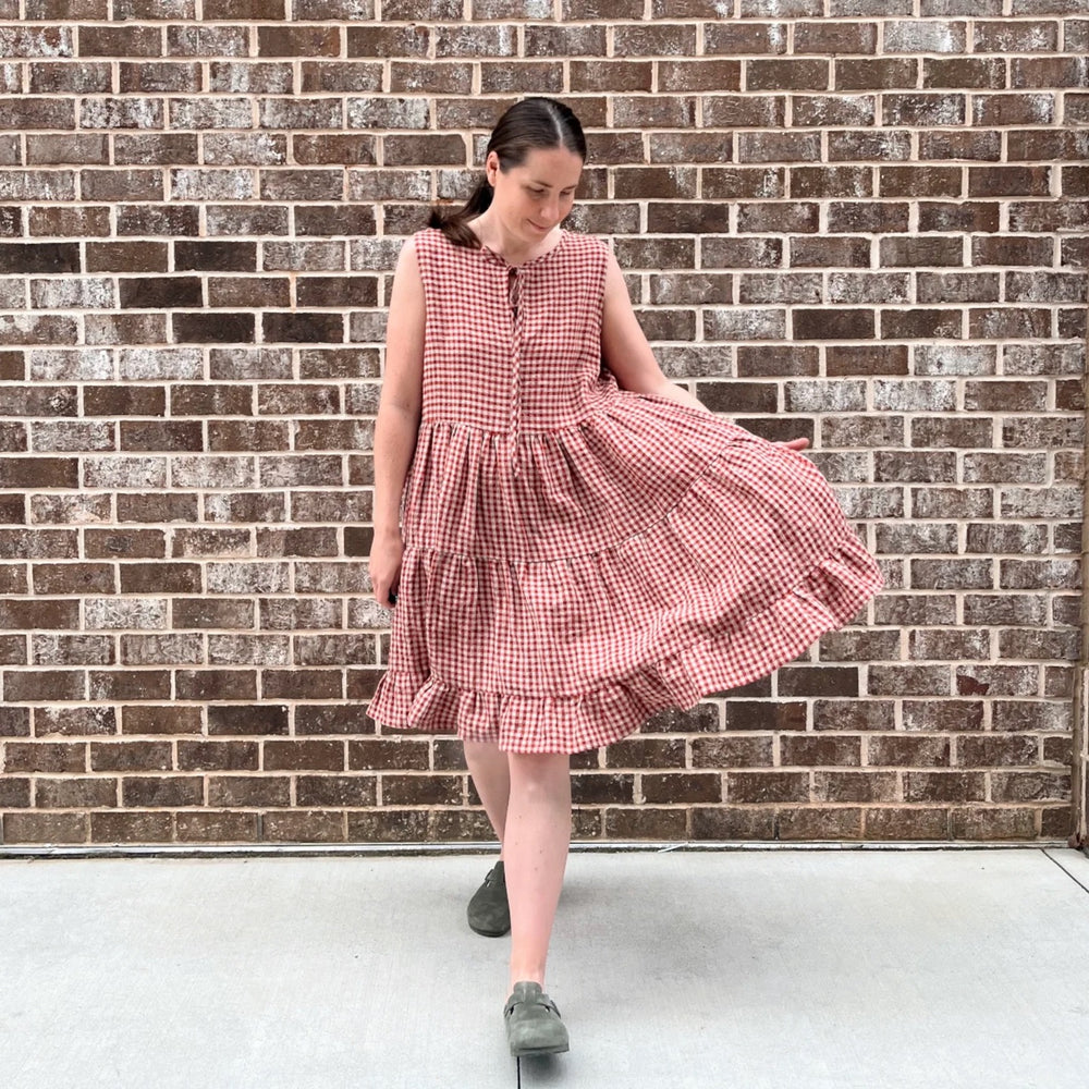 Woman wearing the Babydoll Dress sewing pattern from Me & Kiddo Patterns on The Fold Line. A sleeveless dress pattern made in cotton or linen fabric, featuring a tie at the neckline, three gathered skirt tiers, pockets, and knee length.