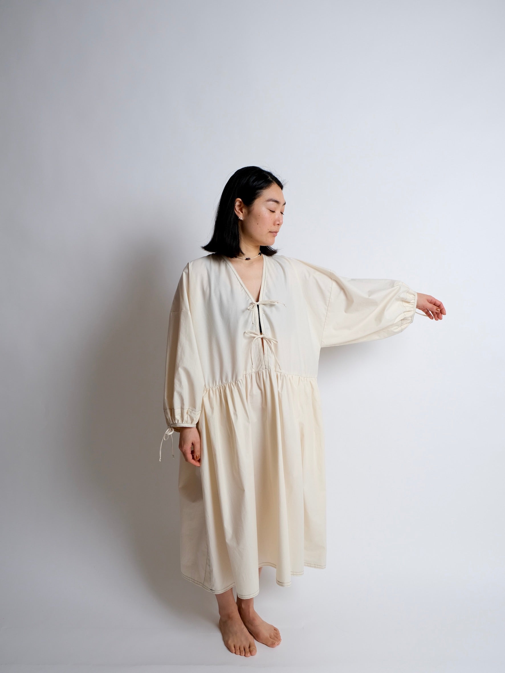 Woman wearing the Bow Dress Hack sewing pattern from Birgitta Helmersson on The Fold Line. An oversized dress pattern made in cotton or linen fabric, featuring a V-neckline, tie front, voluminous long sleeves with ties, a gathered skirt with pockets, and midi length.